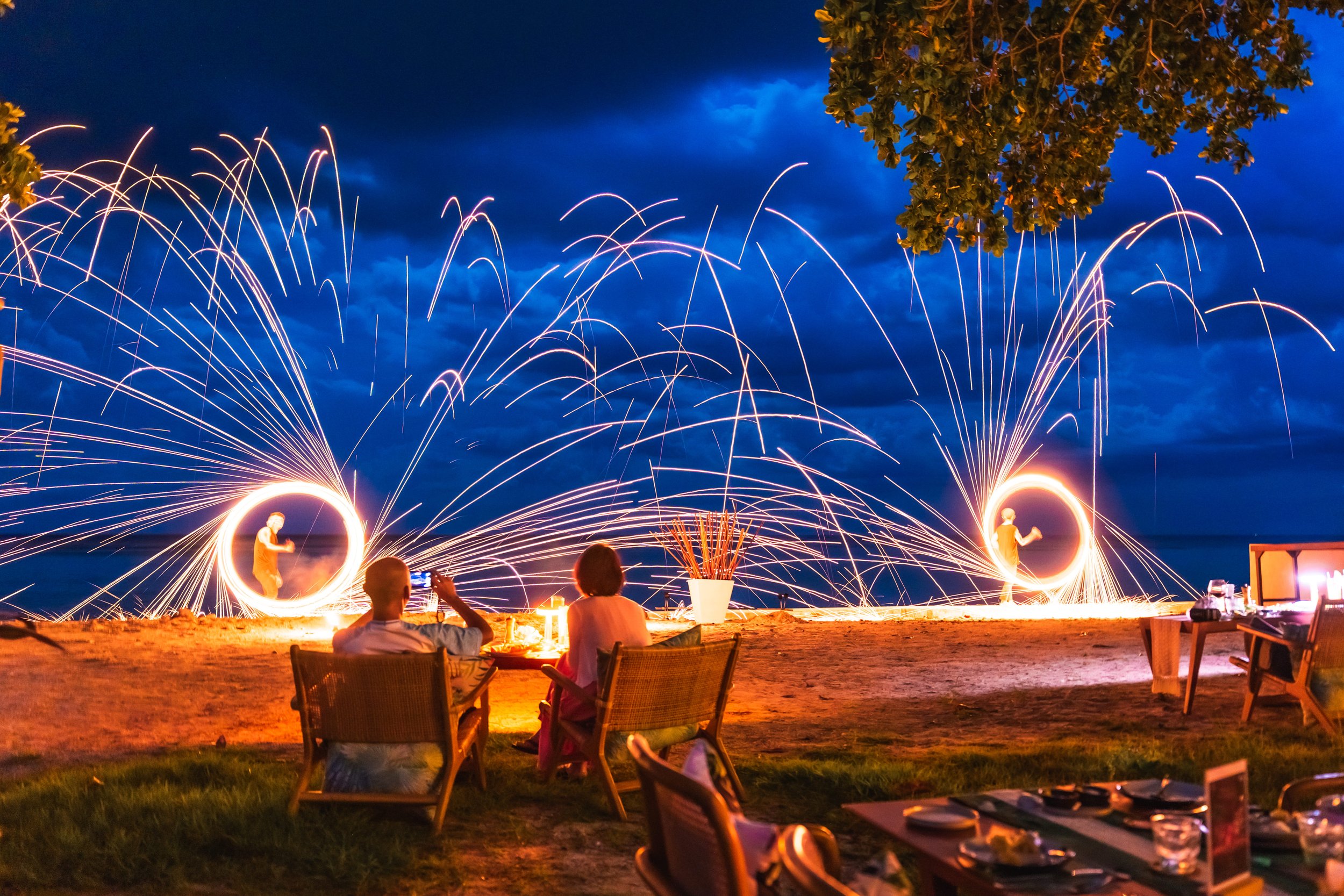 People watching fire spinning show on a beach at night with sparklers and fireworks lighting up the sky.
