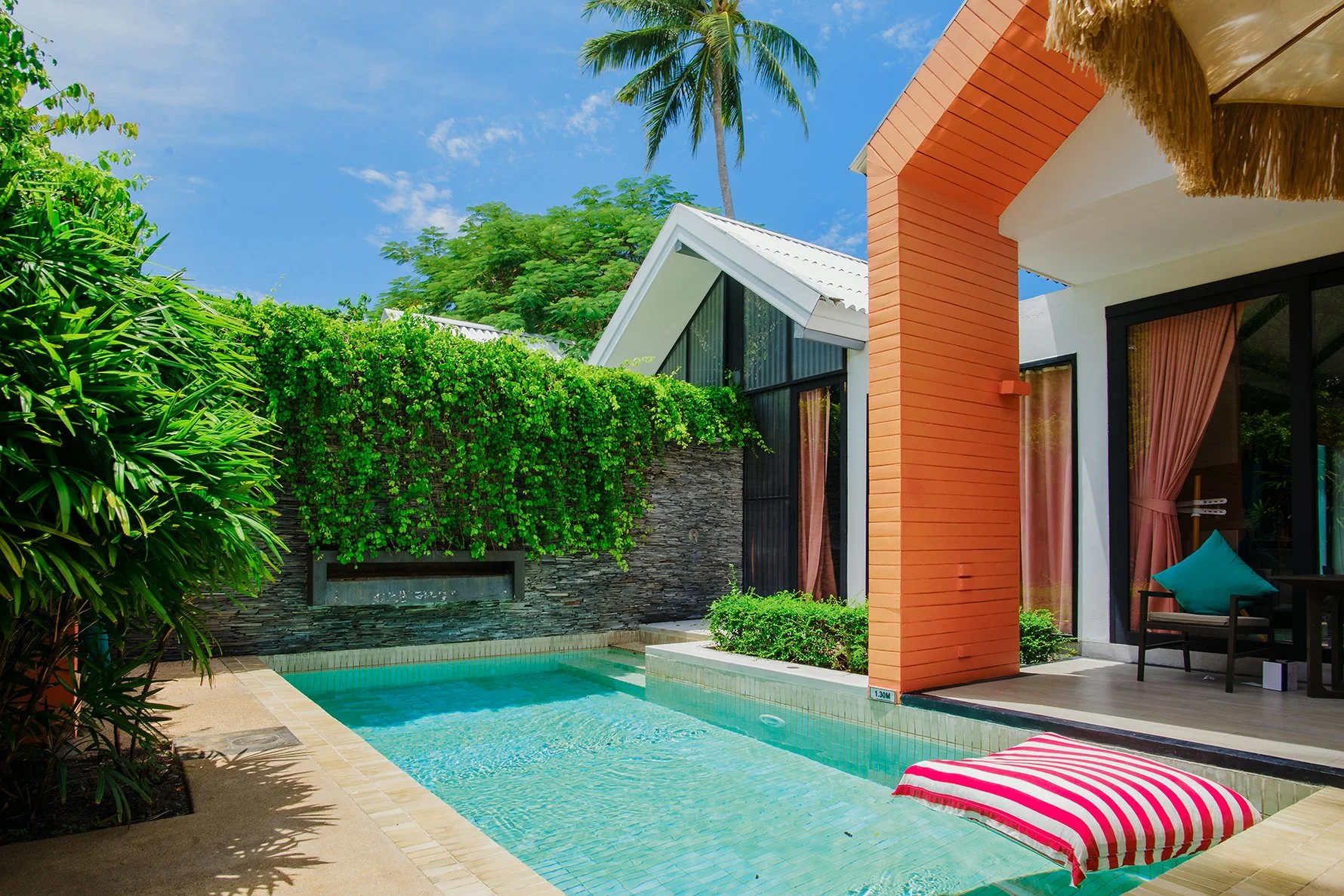 Modern house with a swimming pool, lush green plants, a tall palm tree, a combo of white and orange walls, large glass doors with pink curtains, and blue sky in a tropical setting.