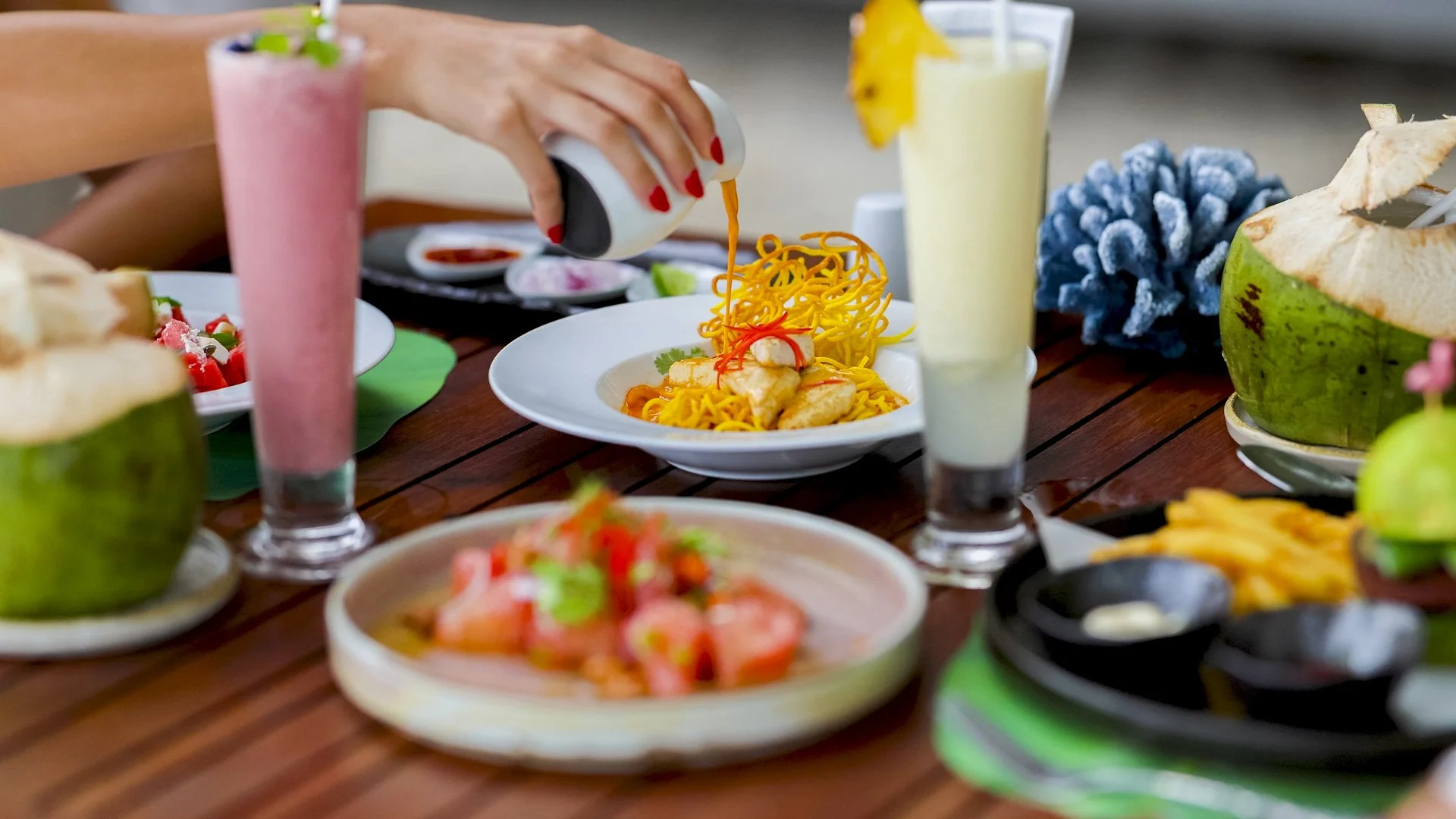 A table set with tropical drinks, dishes of fried chicken and noodles, a fruit salad, and coconuts, with a person's hand pouring sauce on a plate of noodles.
