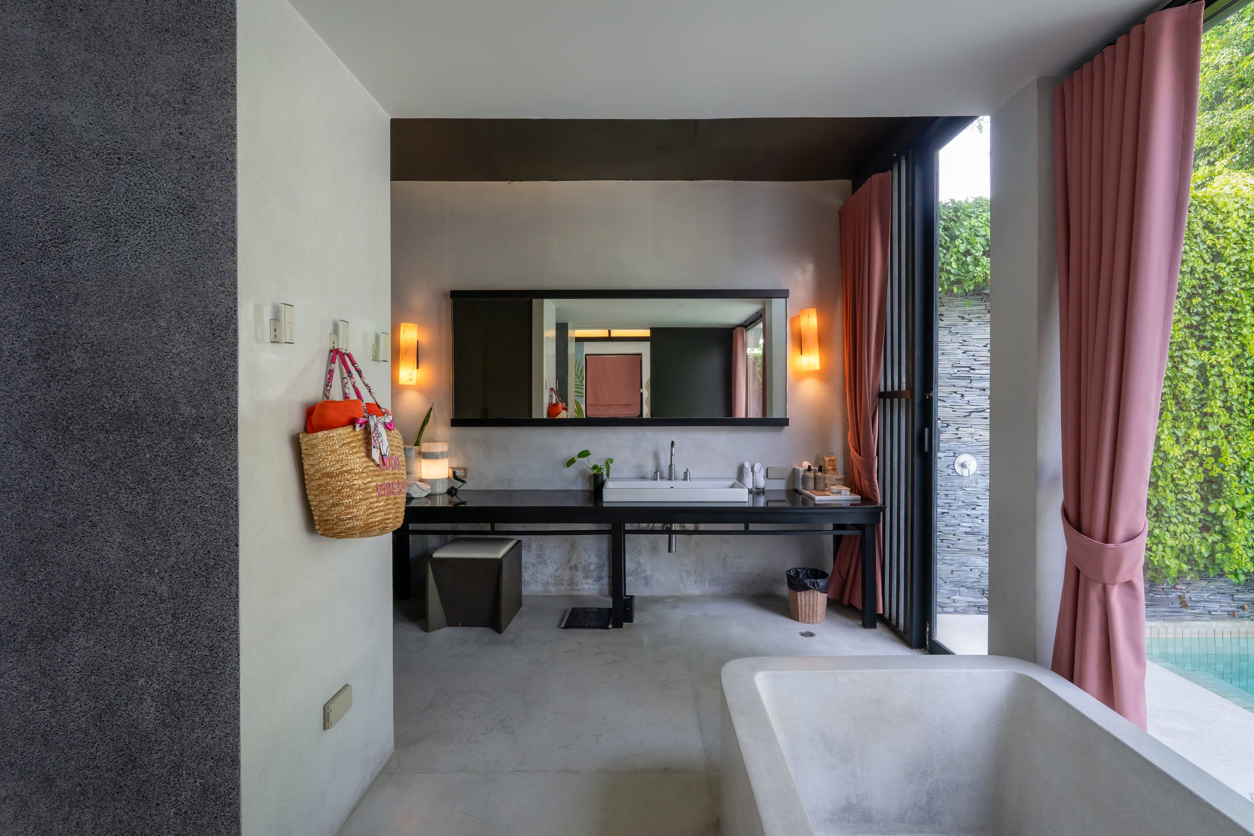 Modern bathroom with a large mirror, beige sink, black counter, pink curtains, and a view of greenery outside.