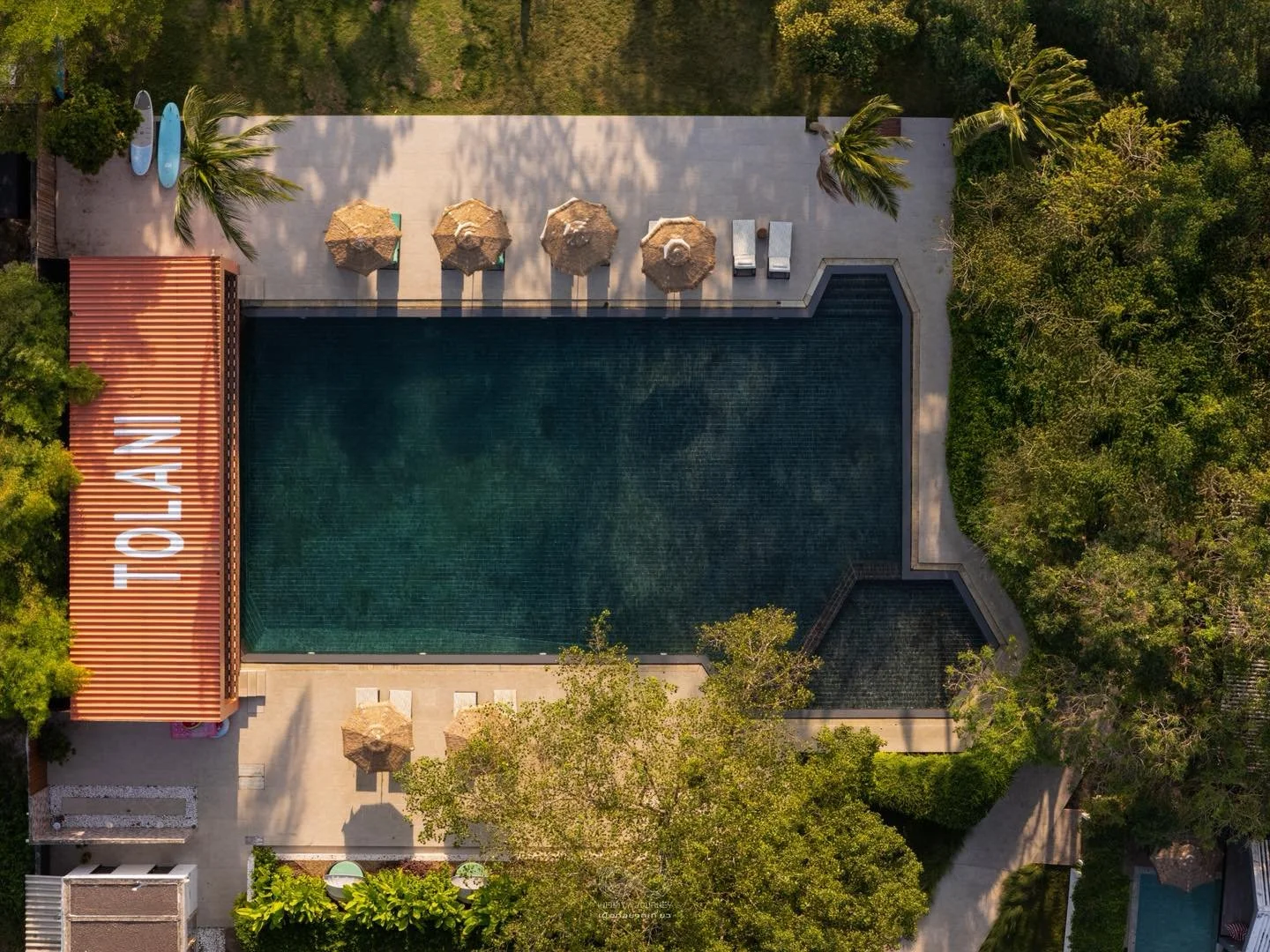 Aerial view of a swimming pool surrounded by trees and a concrete deck, with umbrellas, lounge chairs, and paddleboards. Large 'TOLLAN' sign on a red roofed structure beside the pool.