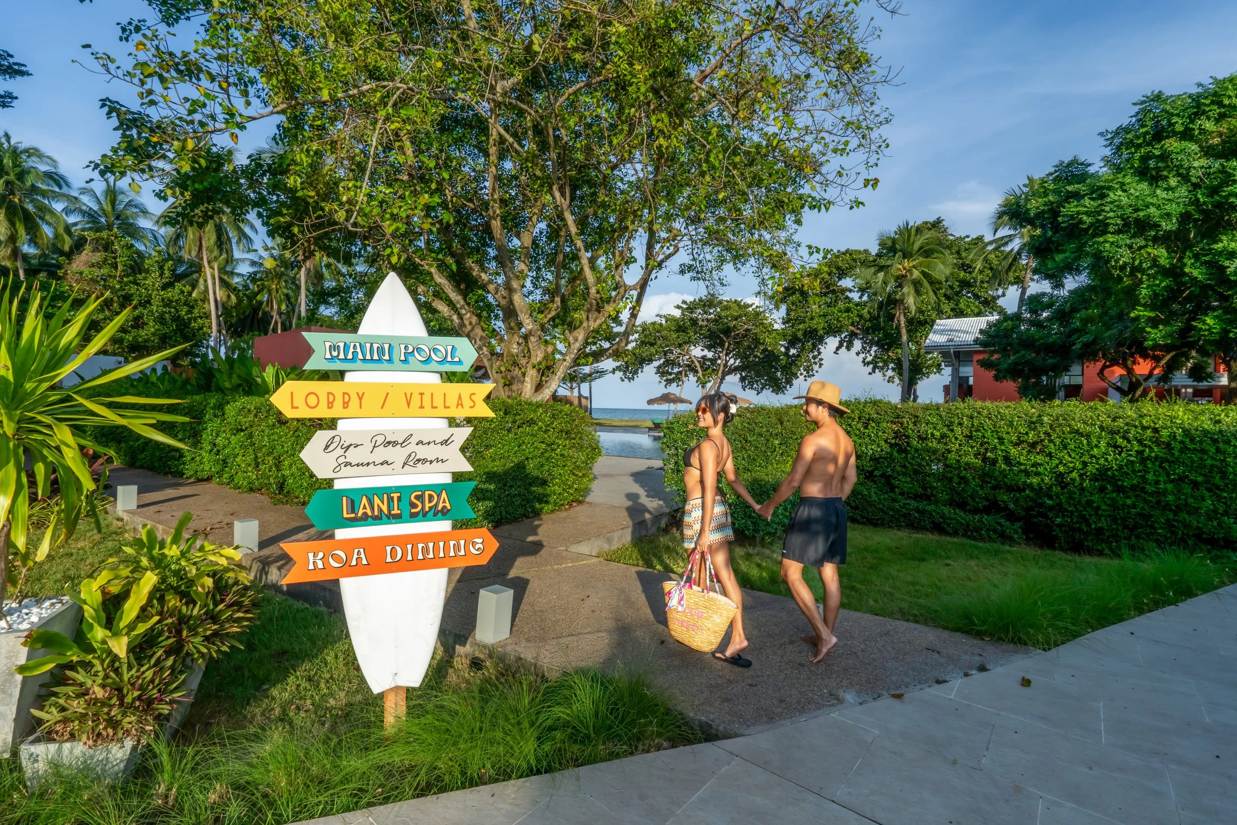 A man and woman holding hands walking near a signpost at a tropical resort with lush greenery, palm trees, a swimming pool, and a glimpse of the ocean in the background.