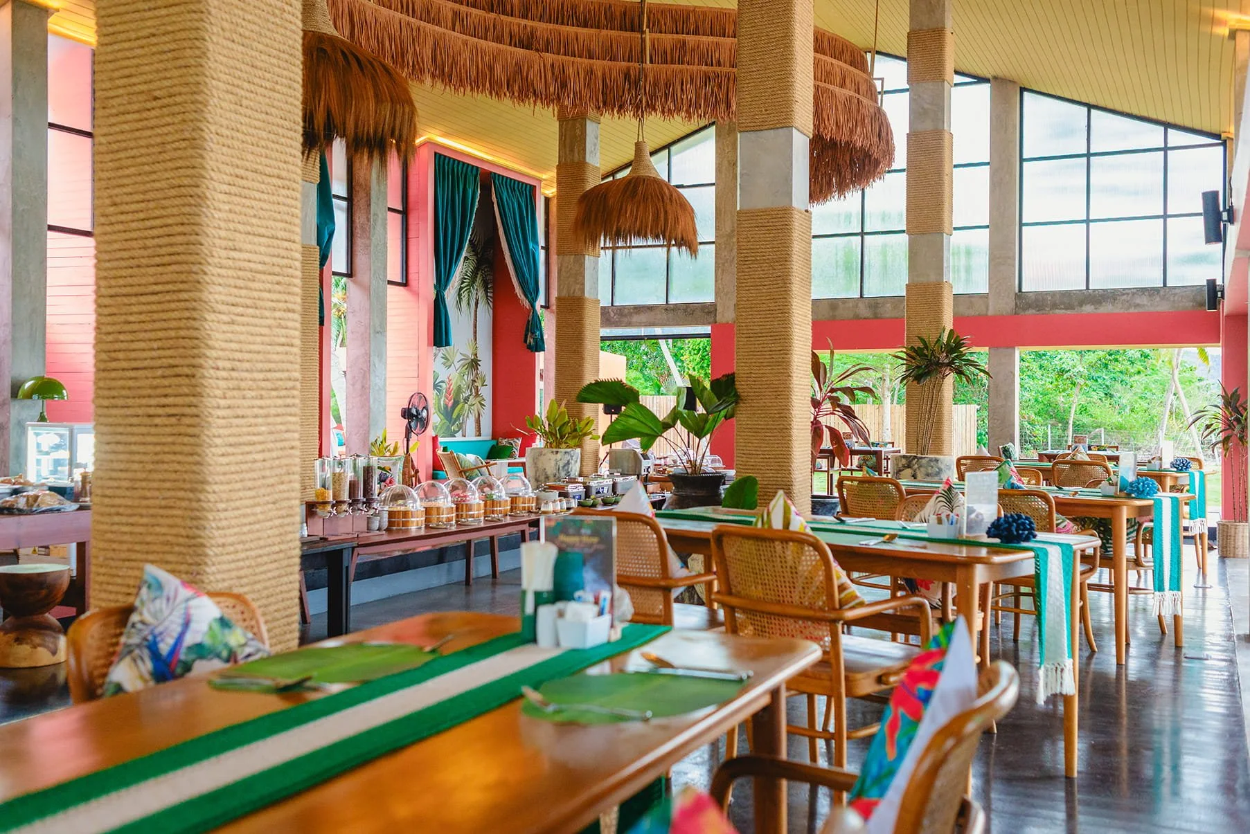 Bright, tropical-themed restaurant interior with wooden tables, colorful chairs, potted plants, and large windows letting in natural light.