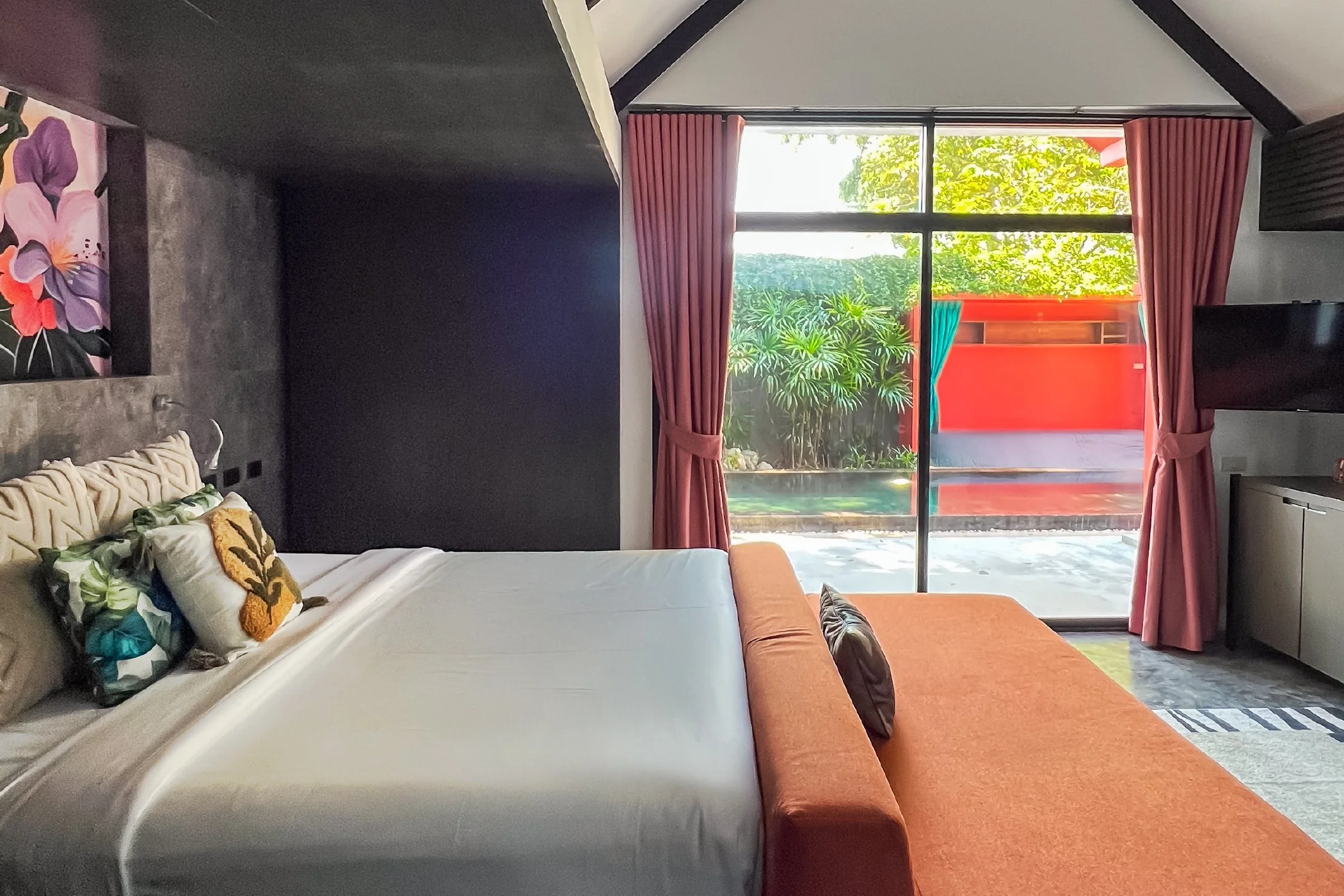 A hotel room with a bed and an orange sofa facing a large sliding glass door, pink curtains, and a view of a small outdoor patio with greenery.