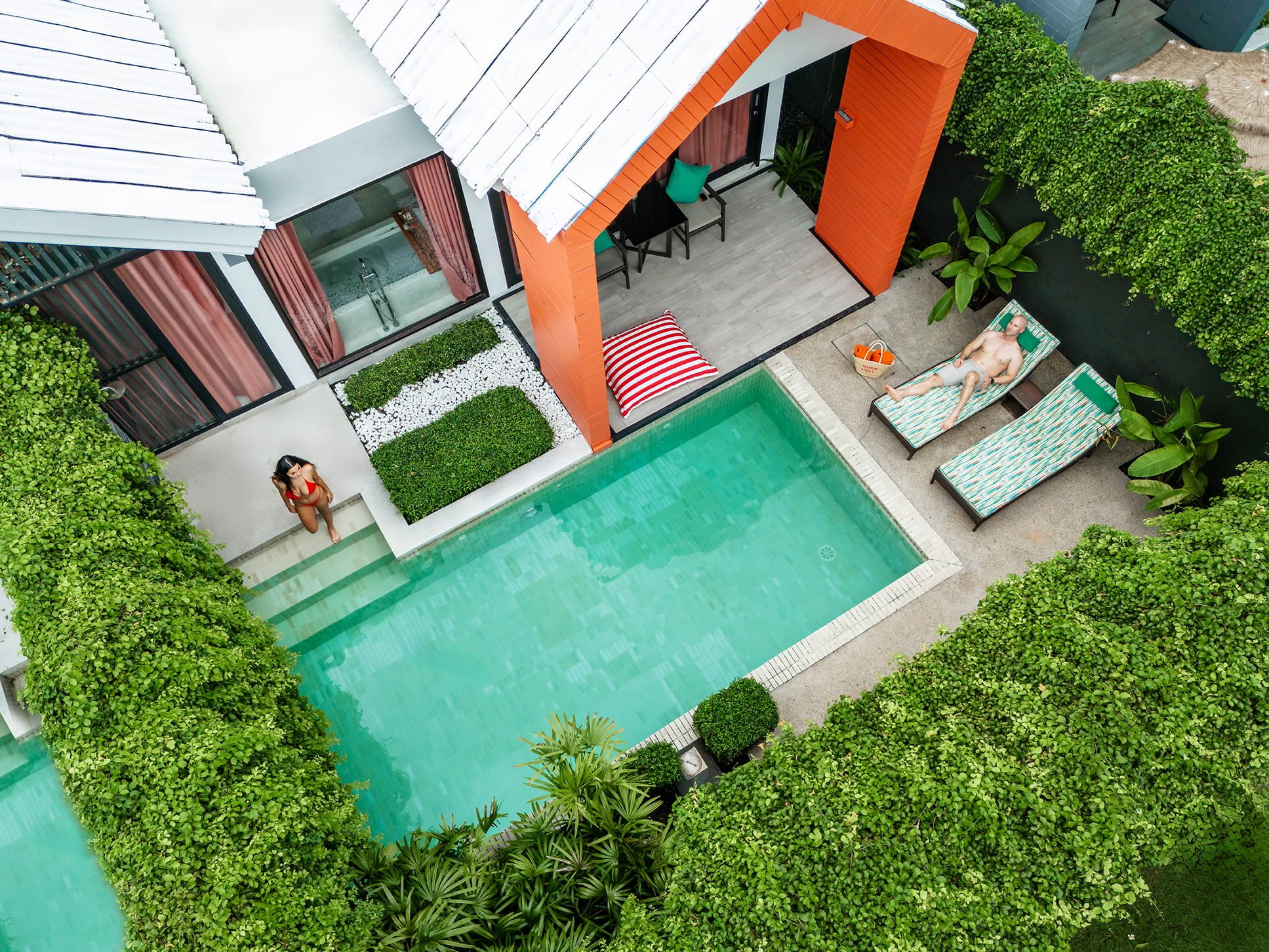 Aerial view of a modern backyard pool area with two people relaxing on lounge chairs, a woman standing near the pool, surrounded by lush green hedges and plants, with parts of a house and a garden visible.