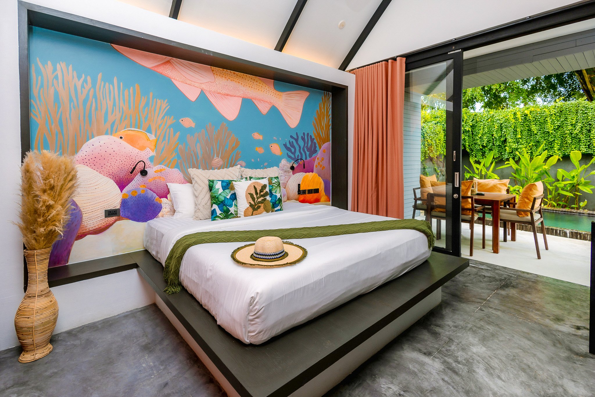 Bedroom with a mural of colorful fish and coral behind the bed. The bed has white bedding, decorative pillows, and a straw hat on top. There is a tall vase with dried plants on the left. Outside the sliding glass door, there is a patio with a table a