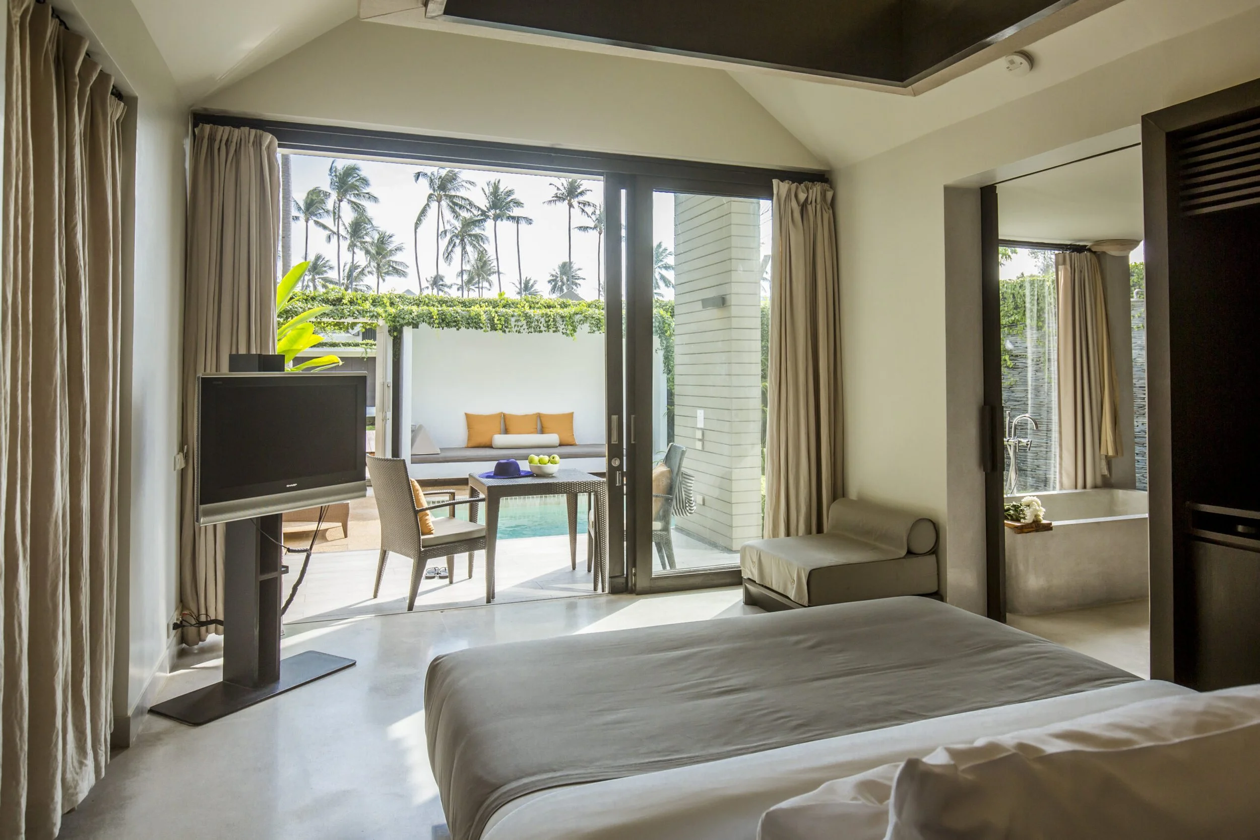 Hotel room with bed, balcony with outdoor seating, pool, and palm trees in the background.