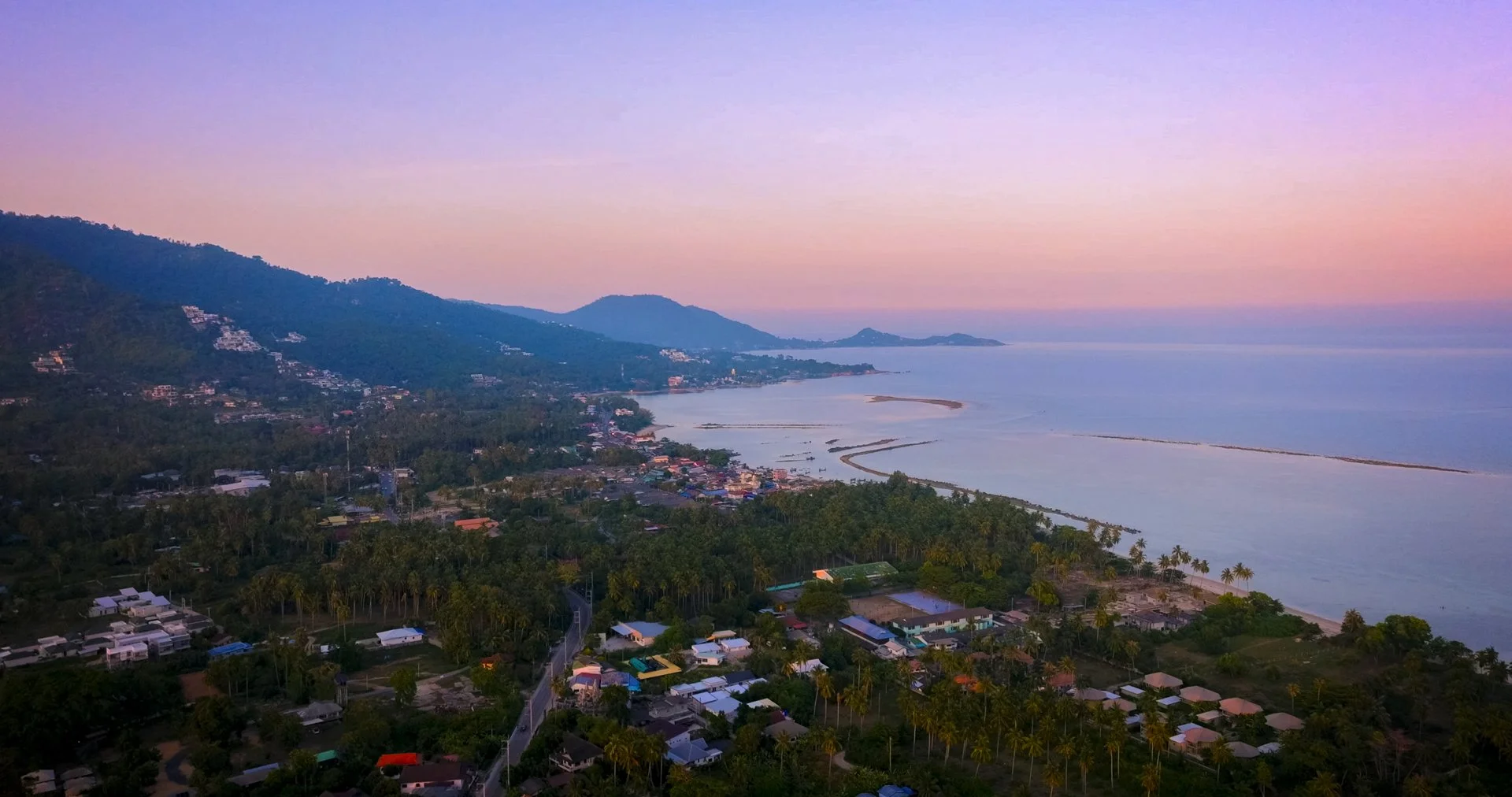 Aerial view of a coastal town with residential houses, trees, and a beach at sunset, with hills in the background and calm ocean waters.