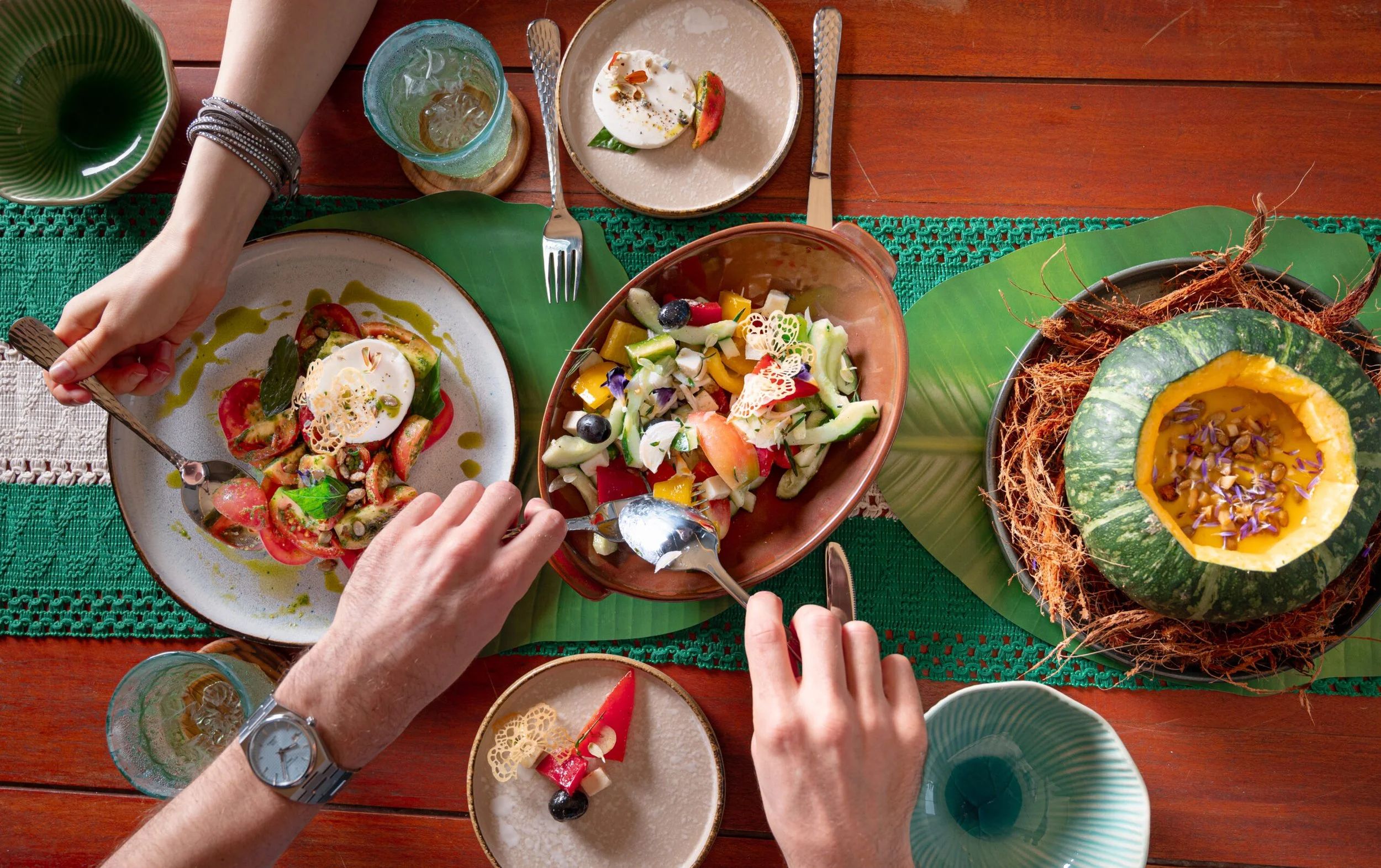 A colorful meal on a wooden table with various salads and a carved pumpkin serving as a bowl for soup, surrounded by glasses of drinks, one person reaching for salad.