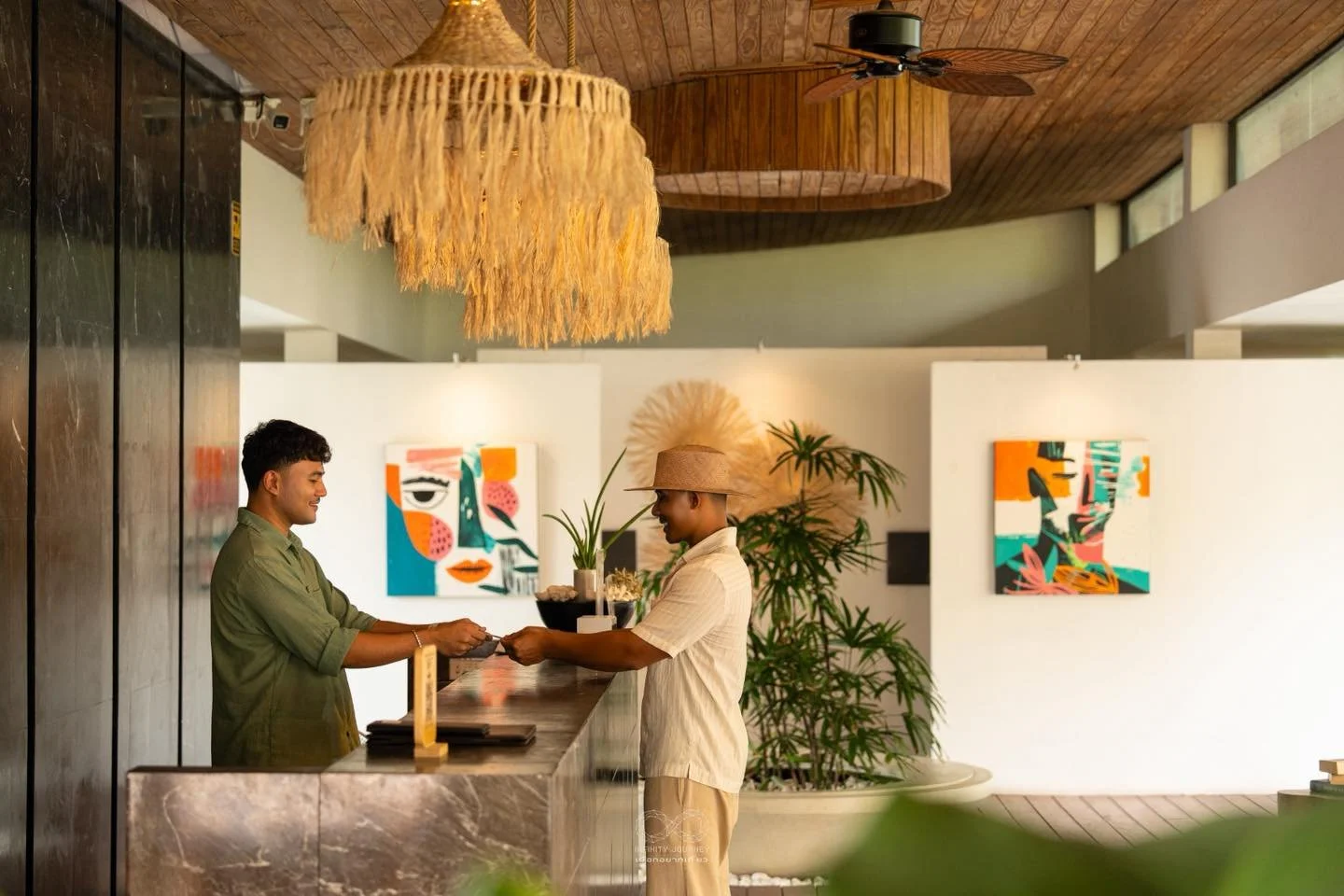 A man at a reception desk in a hotel or resort, handing a keycard or document to a woman, with colorful abstract artwork and green plants on the wall behind them.