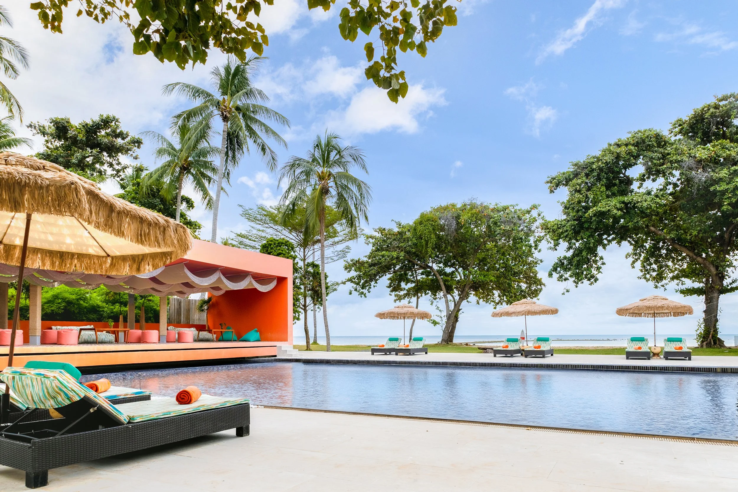 Luxury pool area at a tropical beach resort with lounge chairs, umbrellas, palm trees, and a swimming pool under a partly cloudy sky.