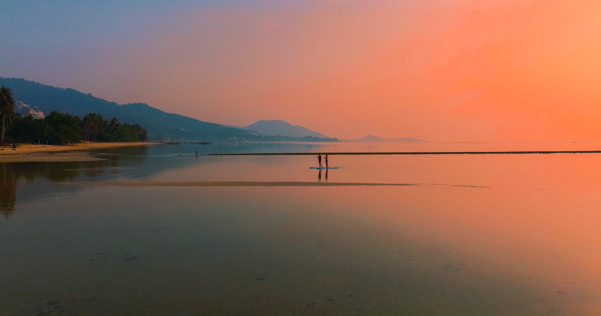 A serene beach during sunset with calm water reflecting the colorful sky, two people on a paddleboard near the shore, and distant mountains covered in trees.