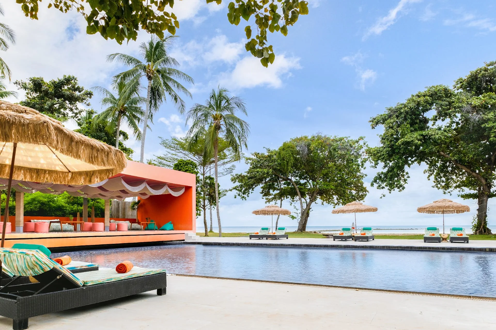 A tropical pool area with sun loungers, beach umbrellas, trees, and a view of the ocean under a partly cloudy sky.