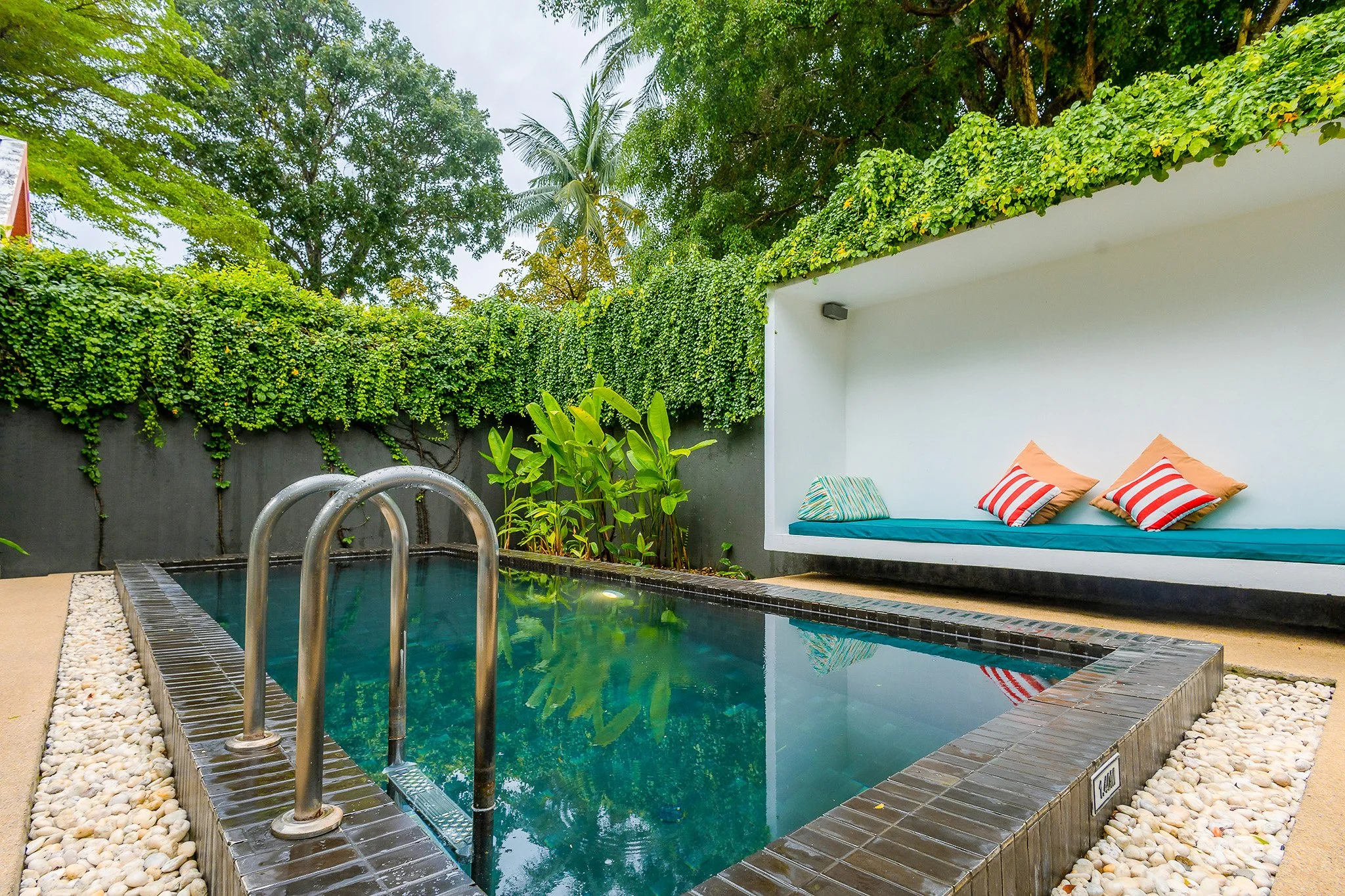 Small private backyard swimming pool with metal handrails, surrounded by pebbles and lush green plants, adjacent to a minimalist white lounge area with cushioned seating and striped pillows, enclosed by a wall with greenery on top.