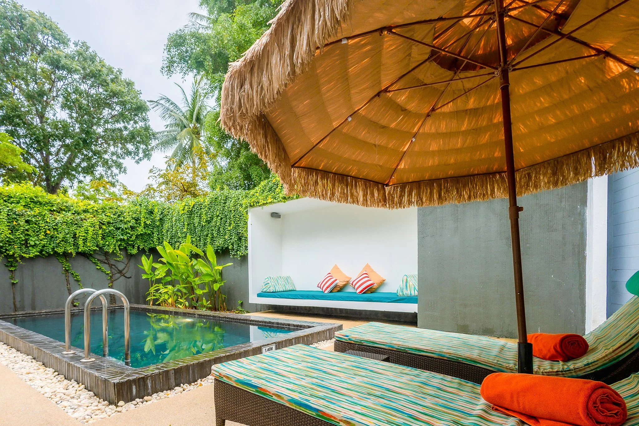 Small swimming pool with metal handrails, surrounded by a beige deck, under a large straw umbrella. There are lounge chairs with striped cushions, a wall with a built-in bench with colorful pillows, and lush green trees and plants in the background.