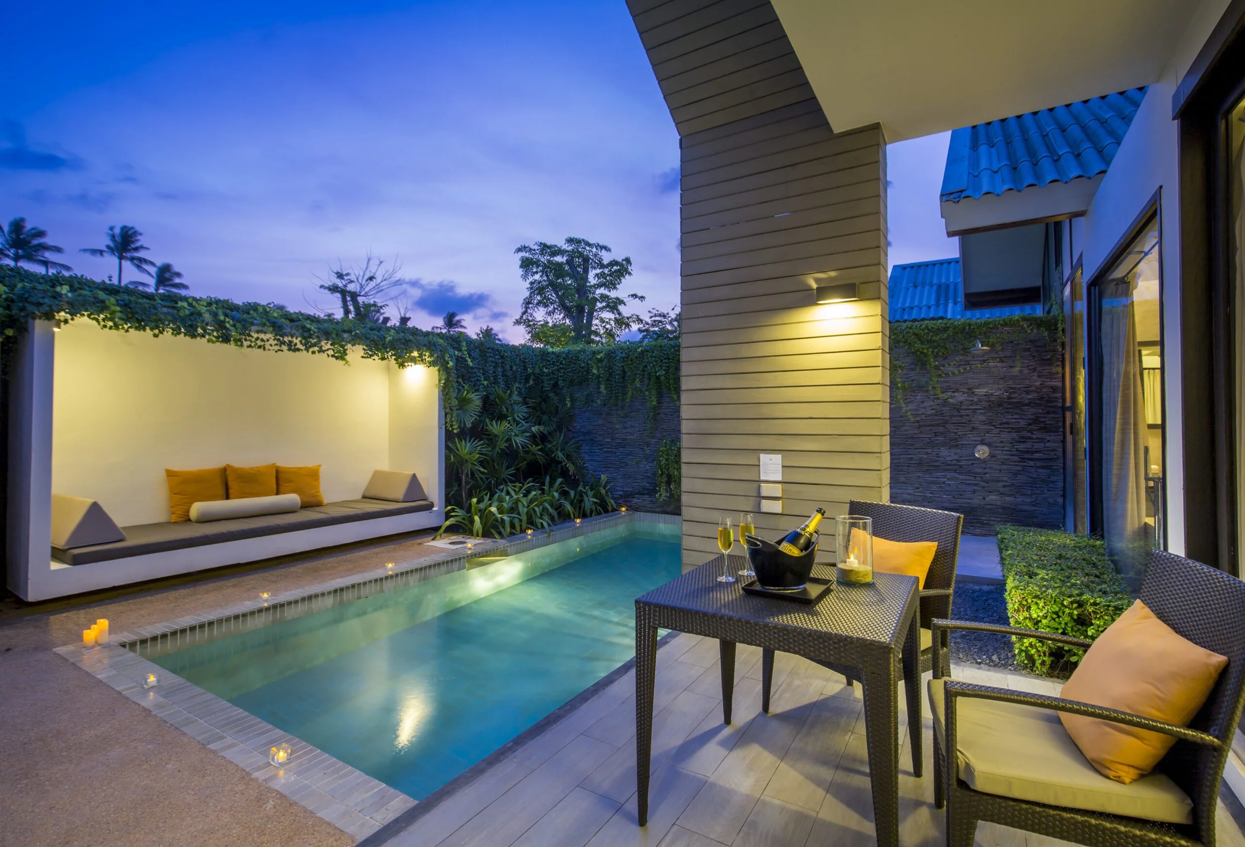 Outdoor patio with a small rectangular pool, lounge seating, and a table with two chairs, set against a darkening evening sky.