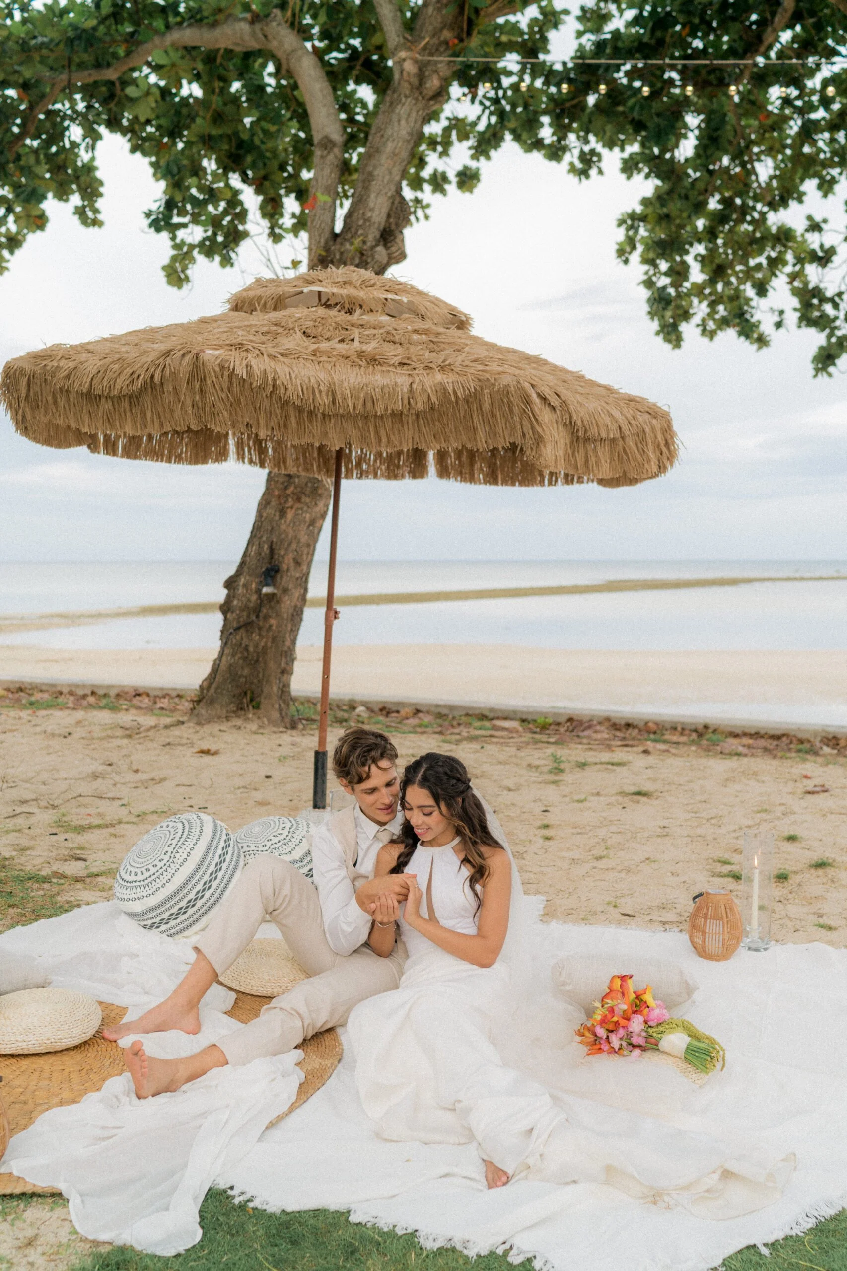 A couple having a beach picnic, sitting on a blanket under a thatched umbrella near the water, with a bouquet of flowers and lit candle nearby.