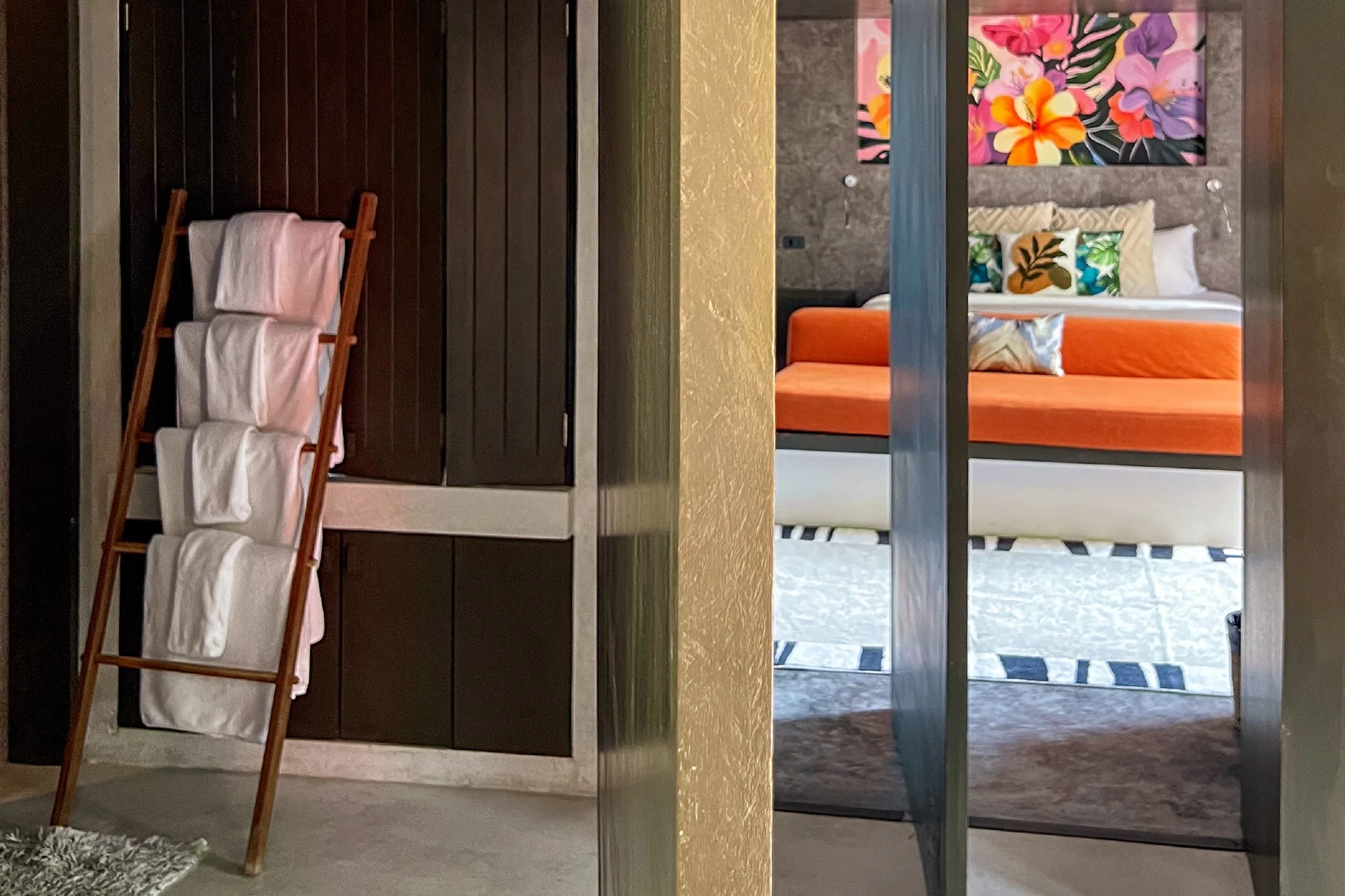 View of a hotel room with a separation wall, showing a bathroom area with toiletries on the left and a bedroom with a bed, colorful artwork, and decorative pillows on the right.