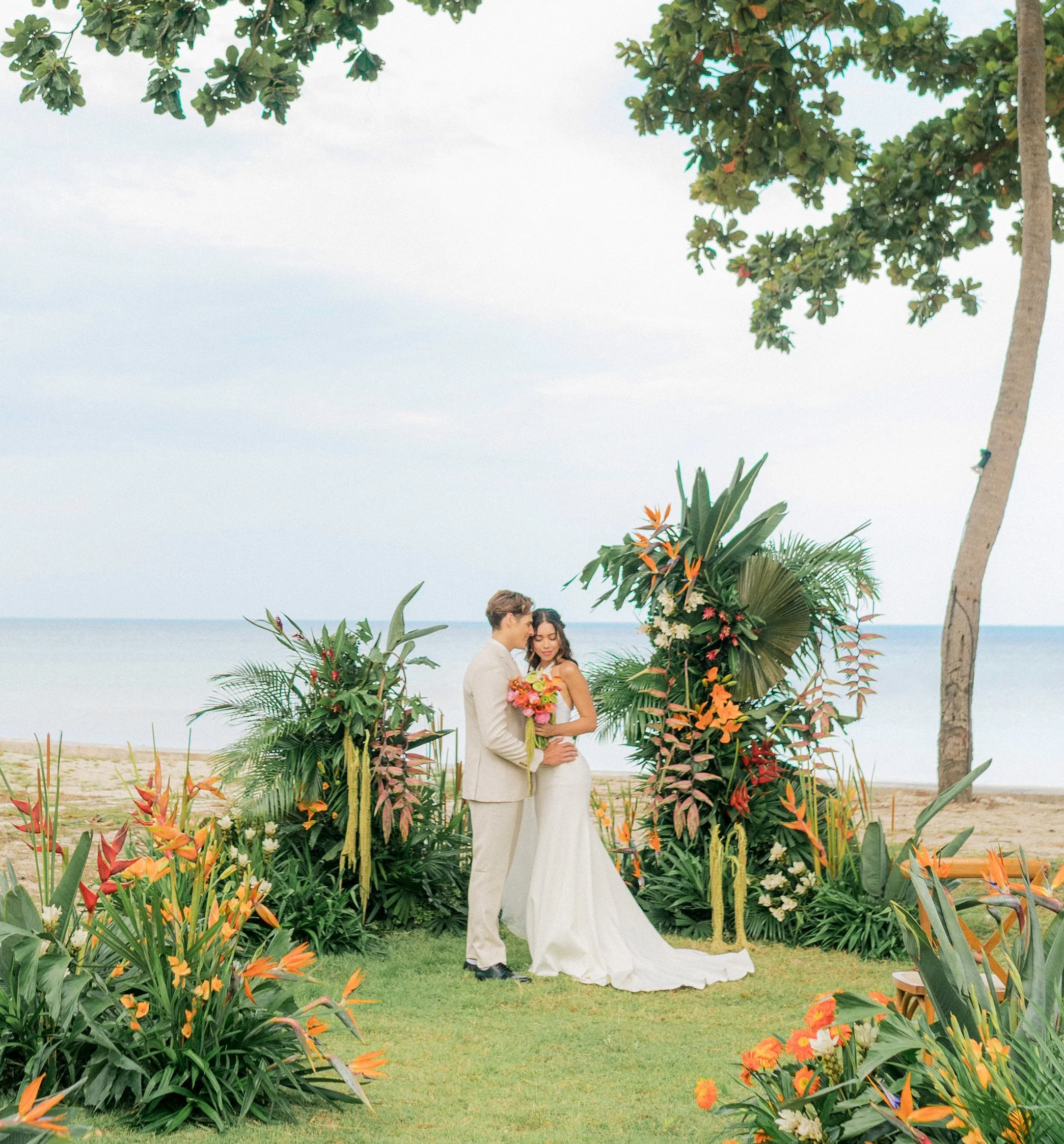 A couple having a beach wedding, standing in front of a floral arrangement with tropical plants, overlooking the ocean with a cloudy sky overhead.