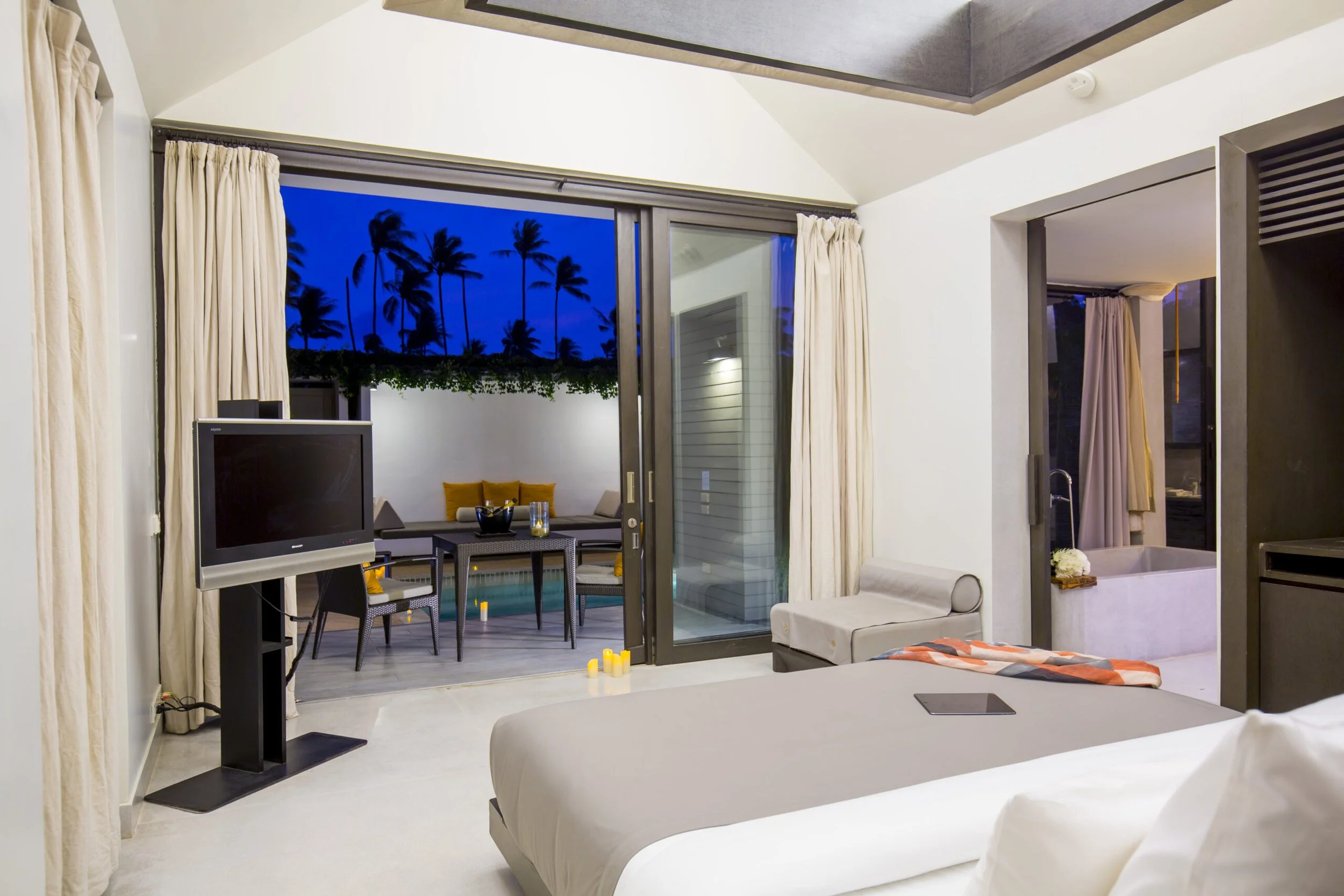 A hotel room with a sliding glass door leading to an outdoor patio with seating and a pool, during nighttime. The room has a white bed, curtains, a TV, and a bathroom visible through an open door.