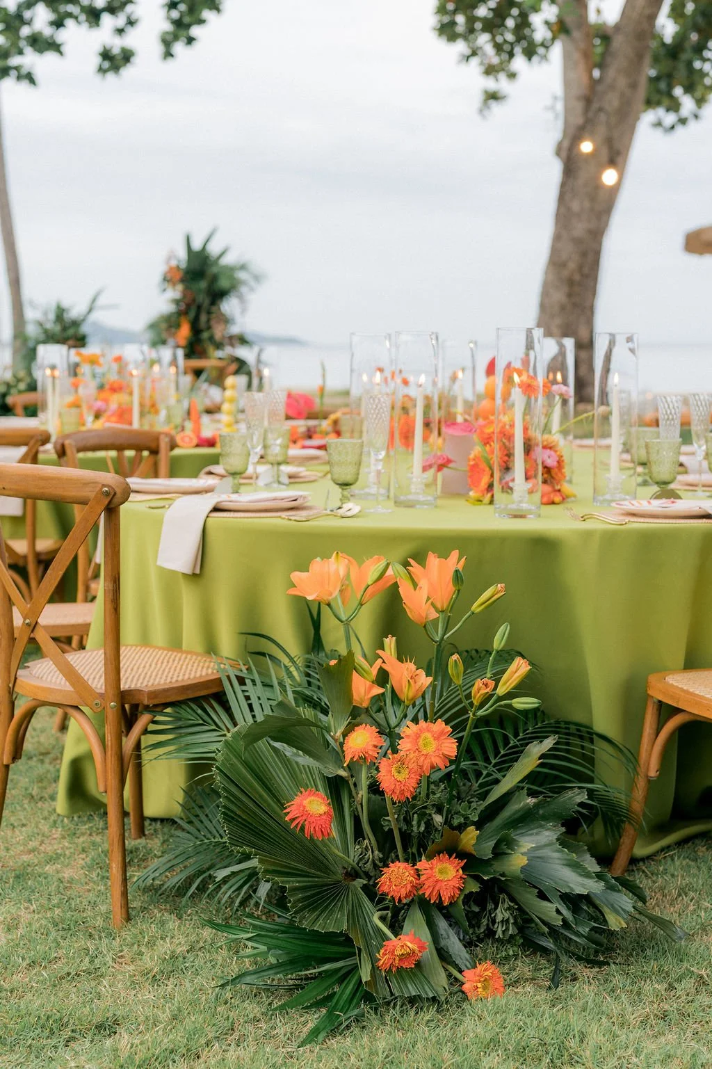 A decorated outdoor event table with a green tablecloth, floral arrangements, and glassware, set against a grassy area and trees with string lights.