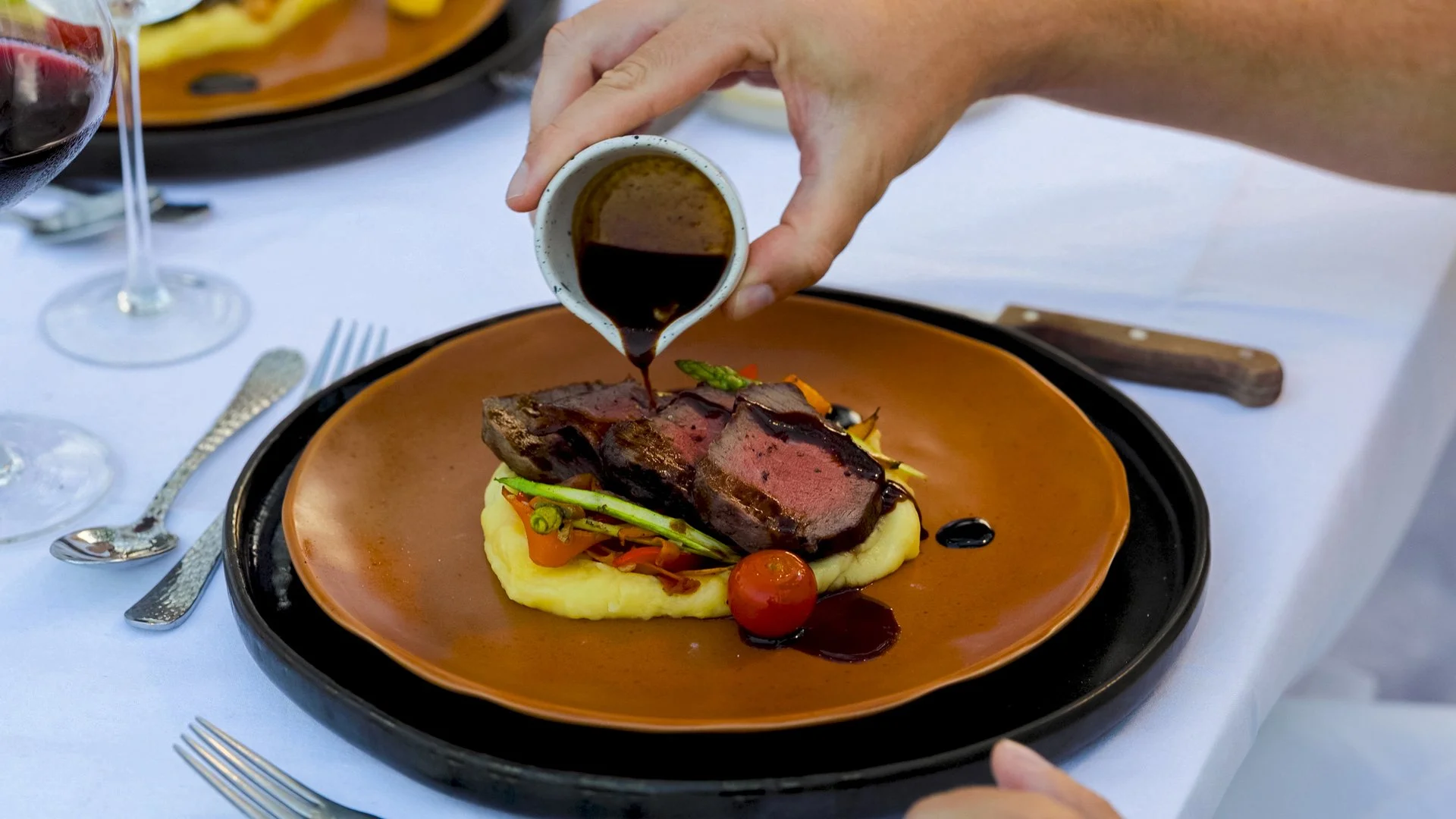 A person pouring sauce over a plated dish of steak, mashed potatoes, vegetables, and cherry tomato at a fine dining restaurant.