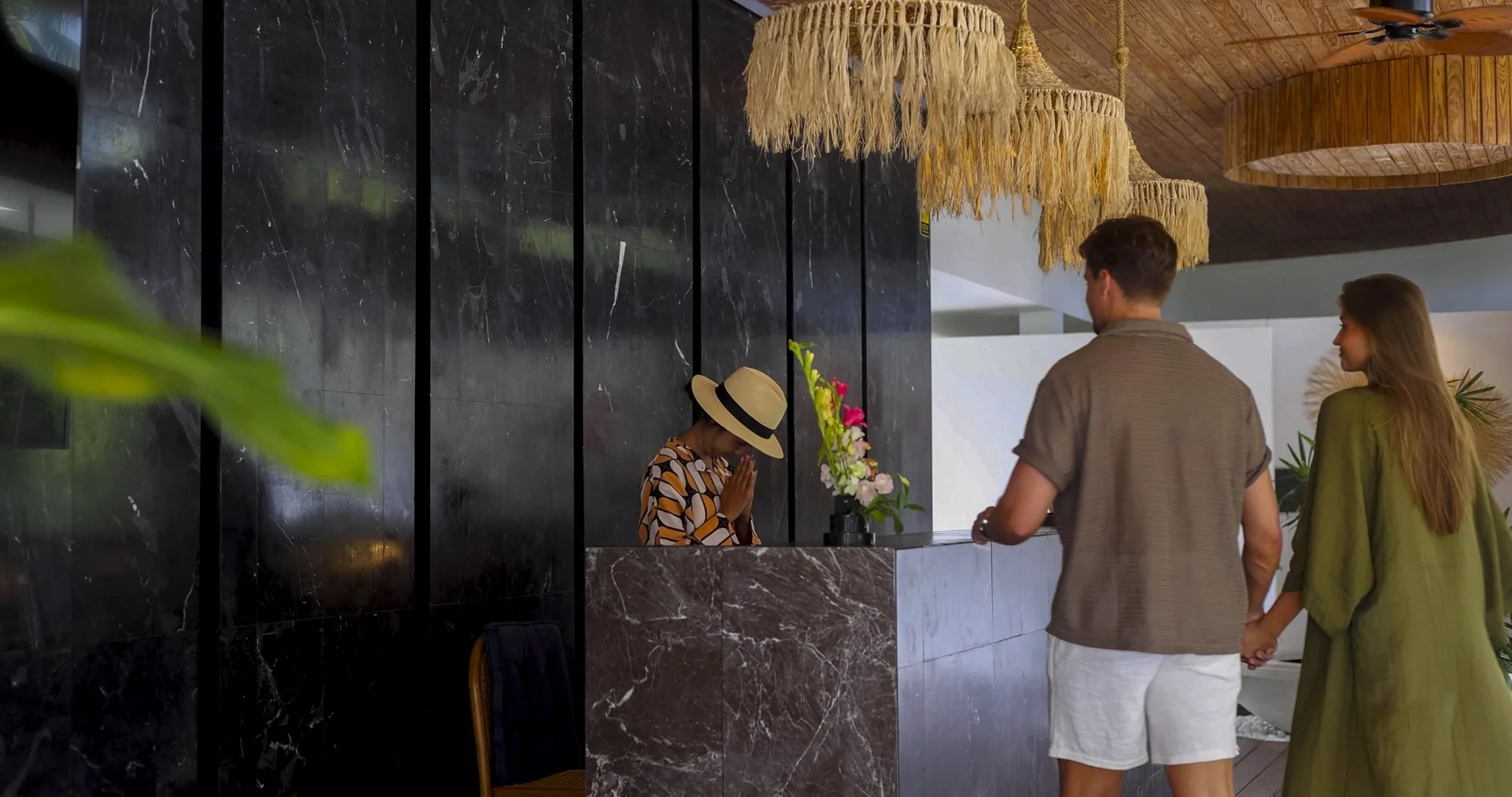 A couple holding hands checks into a hotel reception desk. The receptionist, a woman wearing a hat and patterned shirt, stands behind a dark marble counter with a floral arrangement. The interior has a modern design with hanging woven light fixtures 