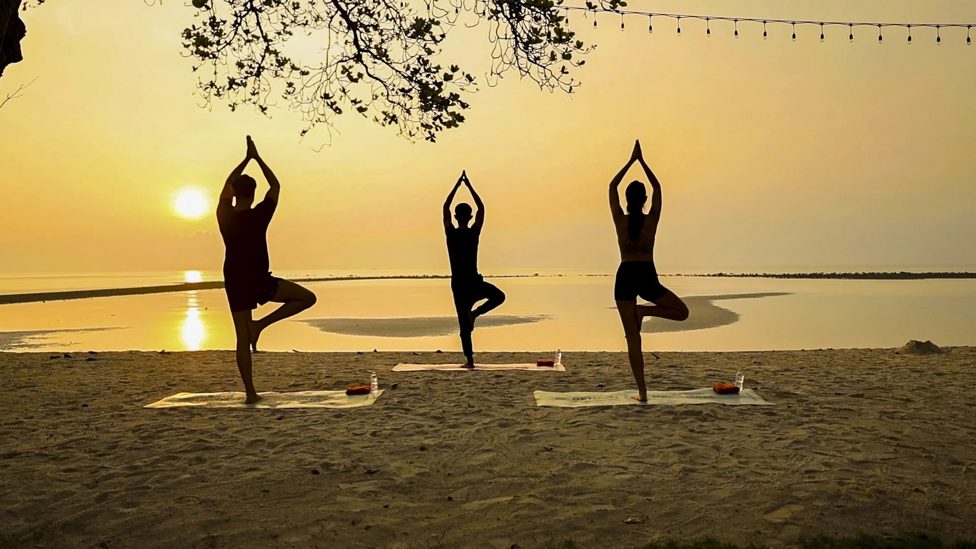Three people practicing yoga on a beach during sunset, standing on one leg with hands together above their heads, looking at the sunset.