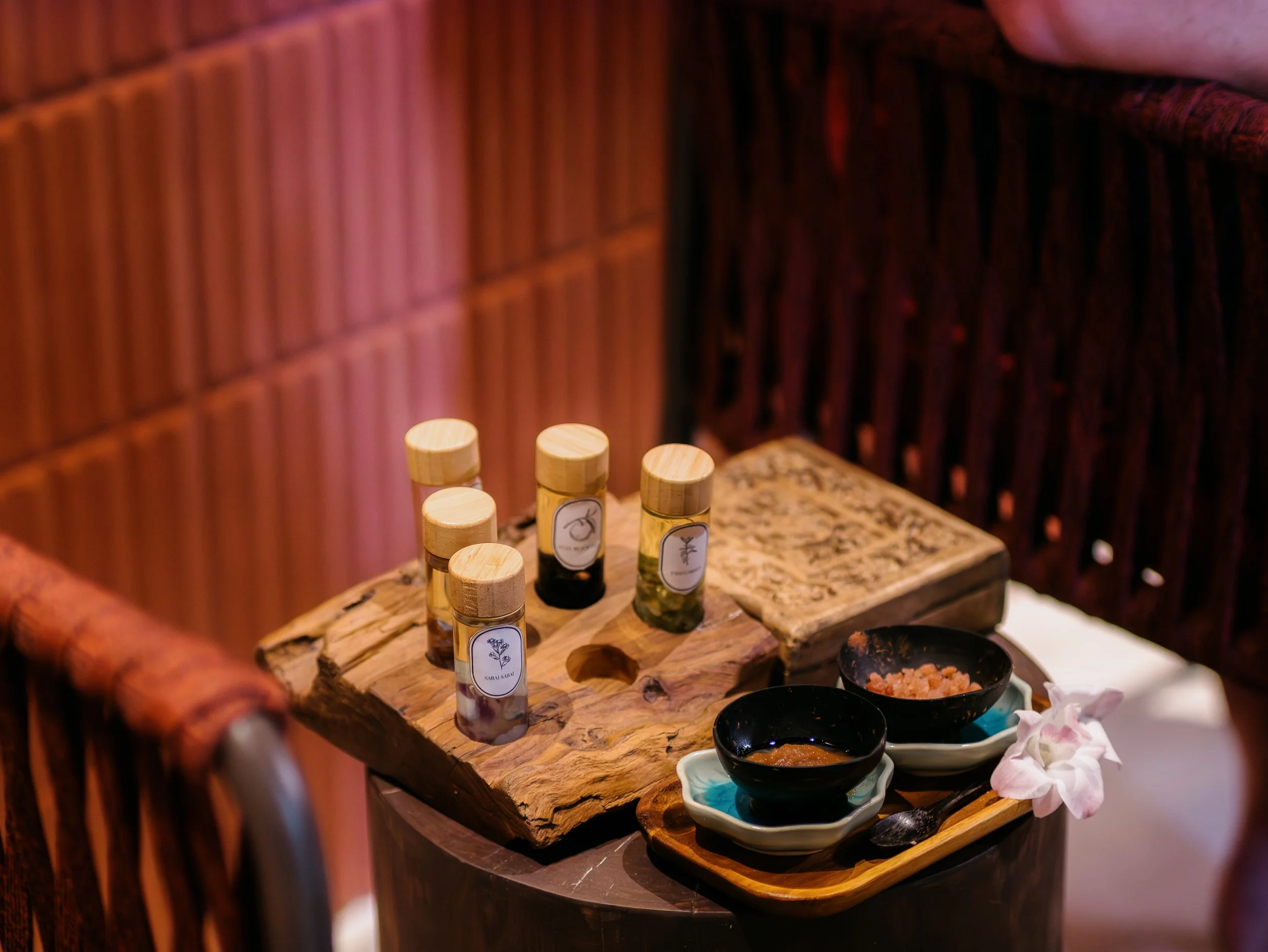 A rustic wooden table set with five small bottles of oil or spices, two black bowls with food, and decorative pink flowers, all situated near bamboo and wooden panels.