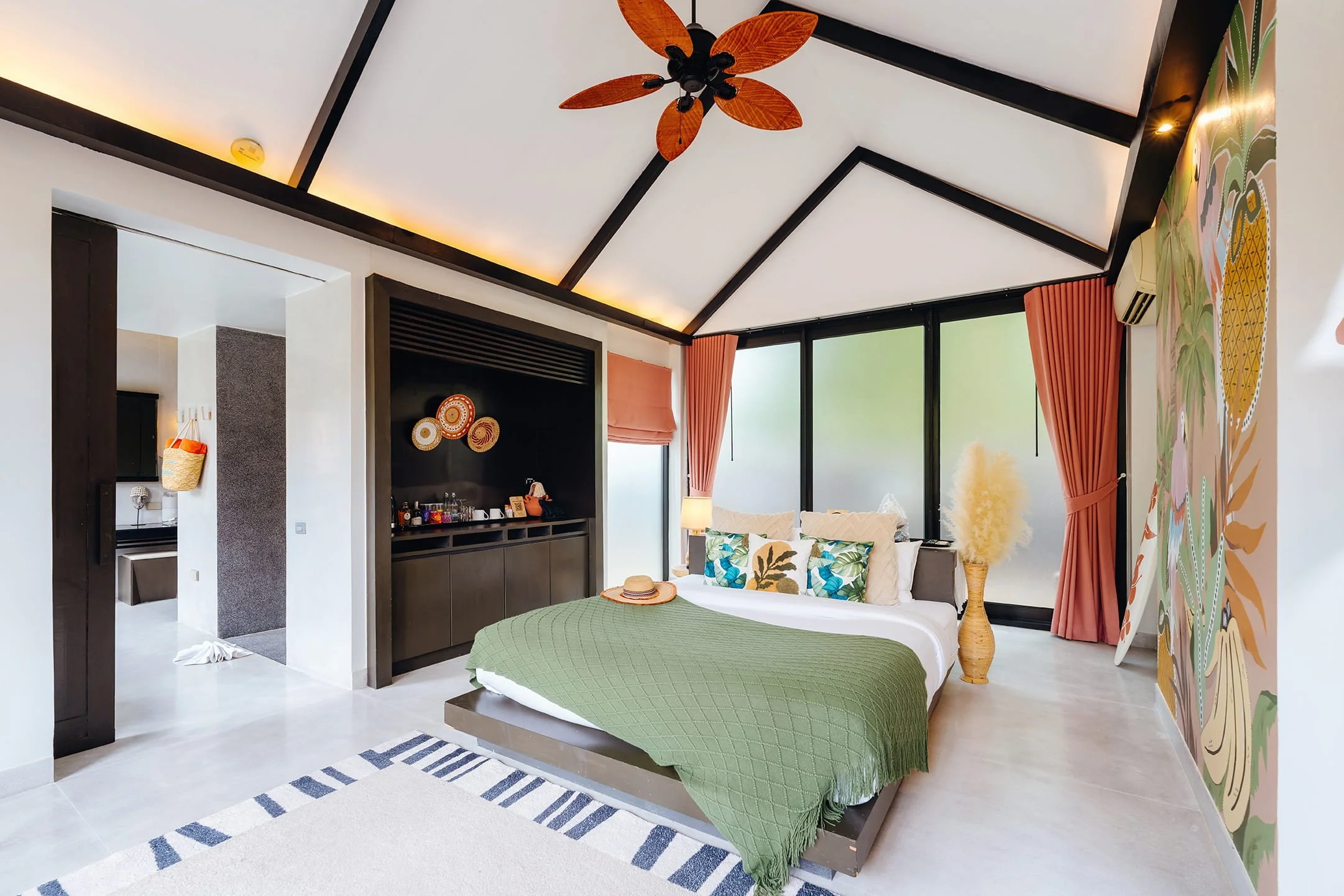 A bright hotel room with a modern design. It features a large bed with white and green bedding, surrounded by decorative pillows, and a straw hat resting on top. The room has large windows with pink curtains, a vase with pampas grass, and colorful wa