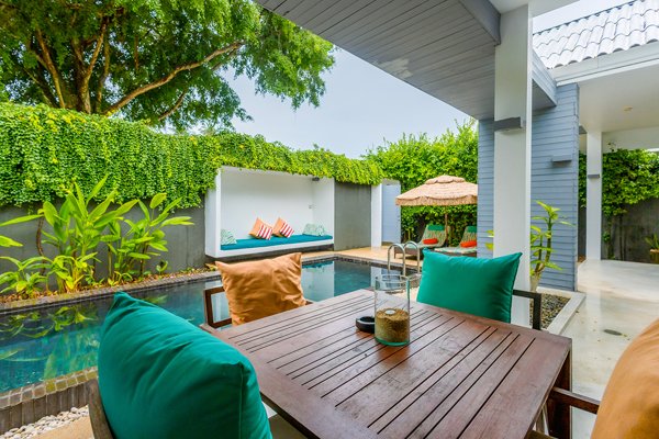 Outdoor patio area with a wooden dining table, colorful cushions, a swimming pool, a shaded lounge area with an umbrella, and lush green plants and trees.