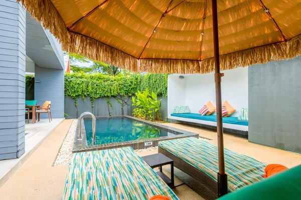 Poolside area with two striped lounge chairs shaded by a large beach umbrella, with a small pool, green plants, and outdoor furniture in the background.