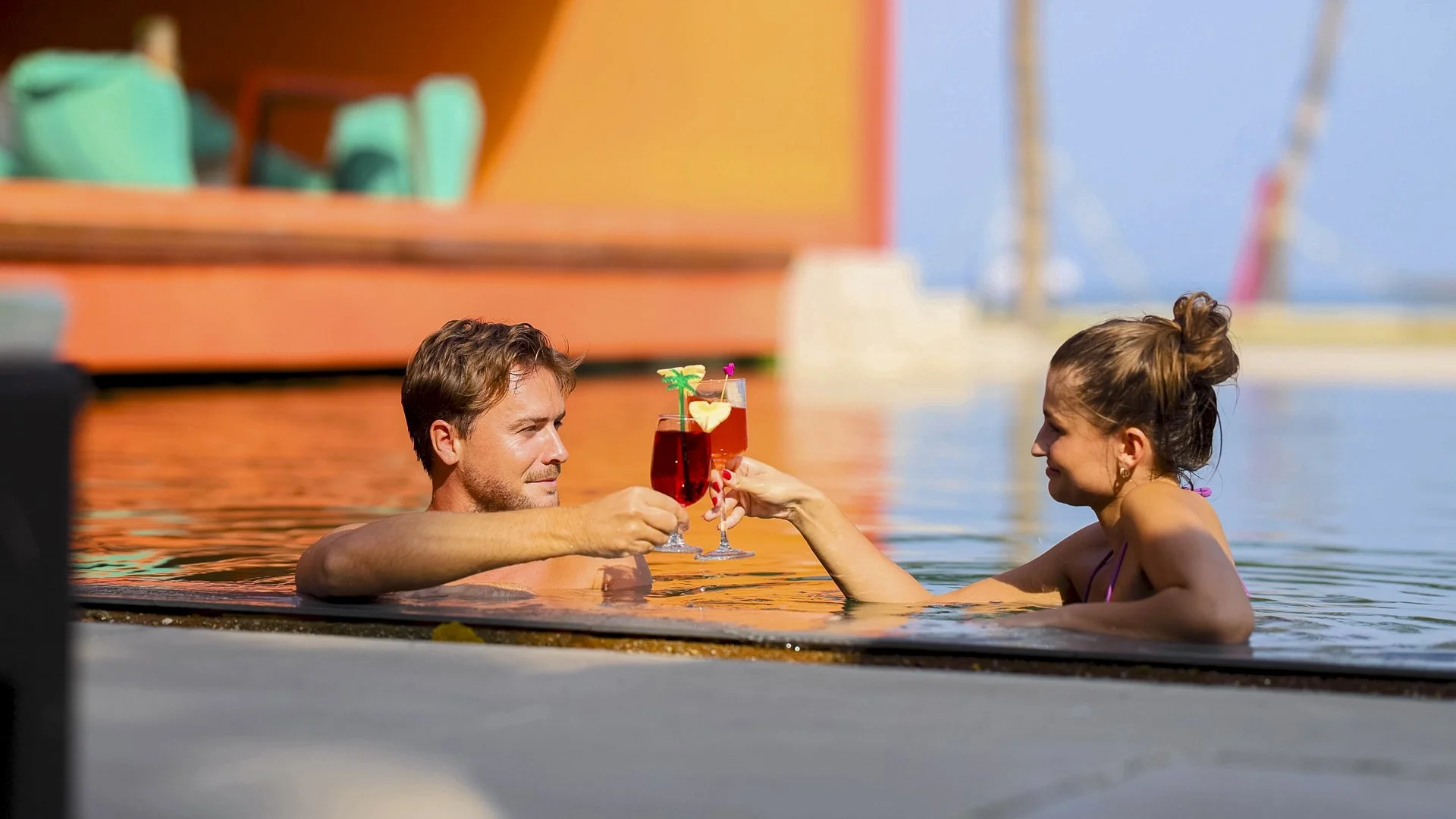 A man and woman in a hot tub outdoors during daytime, clinking glasses with tropical drinks, with a boat and water in the background.