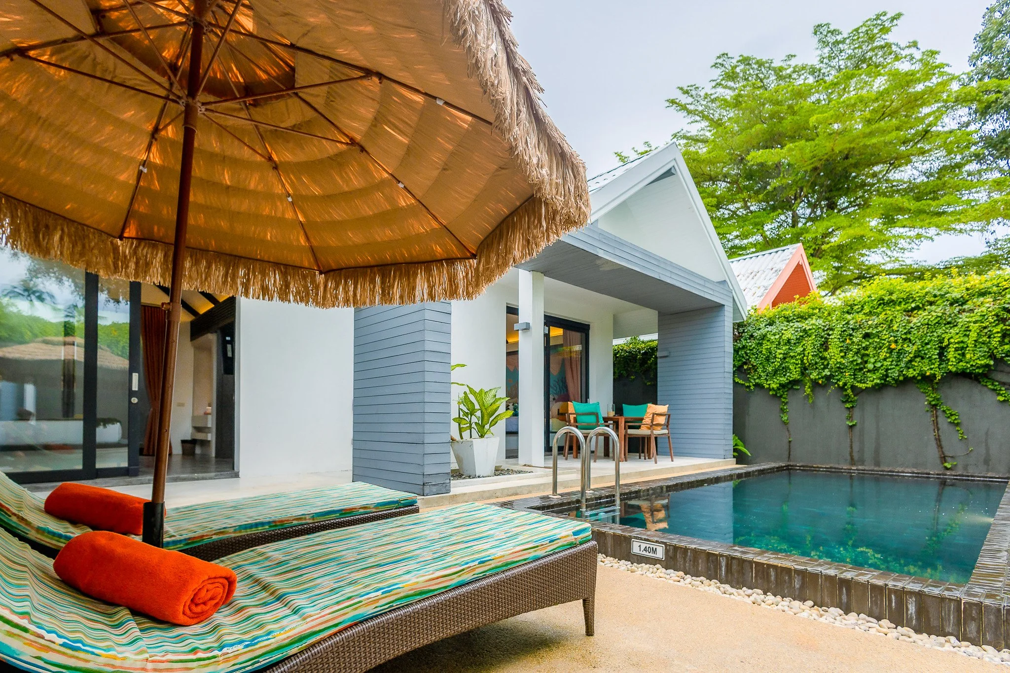 A cozy outdoor pool area with lounge chairs, an orange beach umbrella, and a small seating area next to a modern house with a gray and white exterior, lush green trees, and vines on a wall.