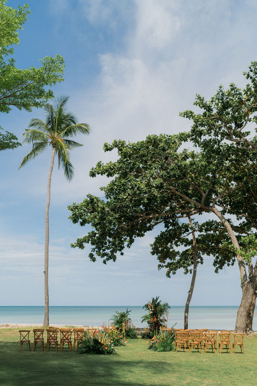 Outdoor beach wedding setup with chairs arranged on green grass, tropical trees, and a view of the ocean under a partly cloudy sky.