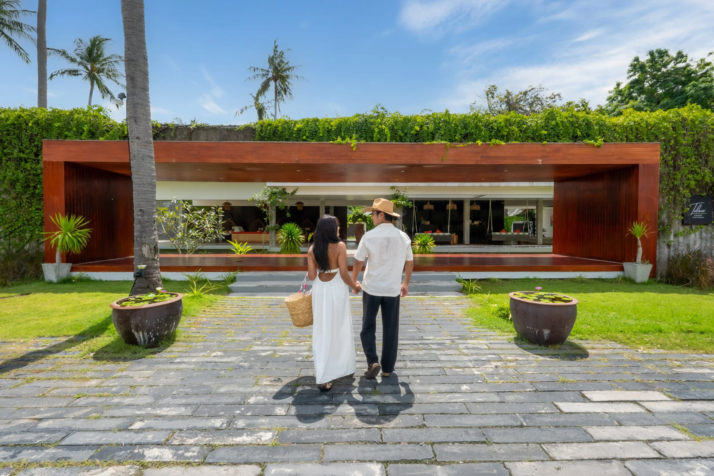 A couple walking hand in hand towards a modern building with a wooden facade, surrounded by greenery and potted plants, on a sunny day.