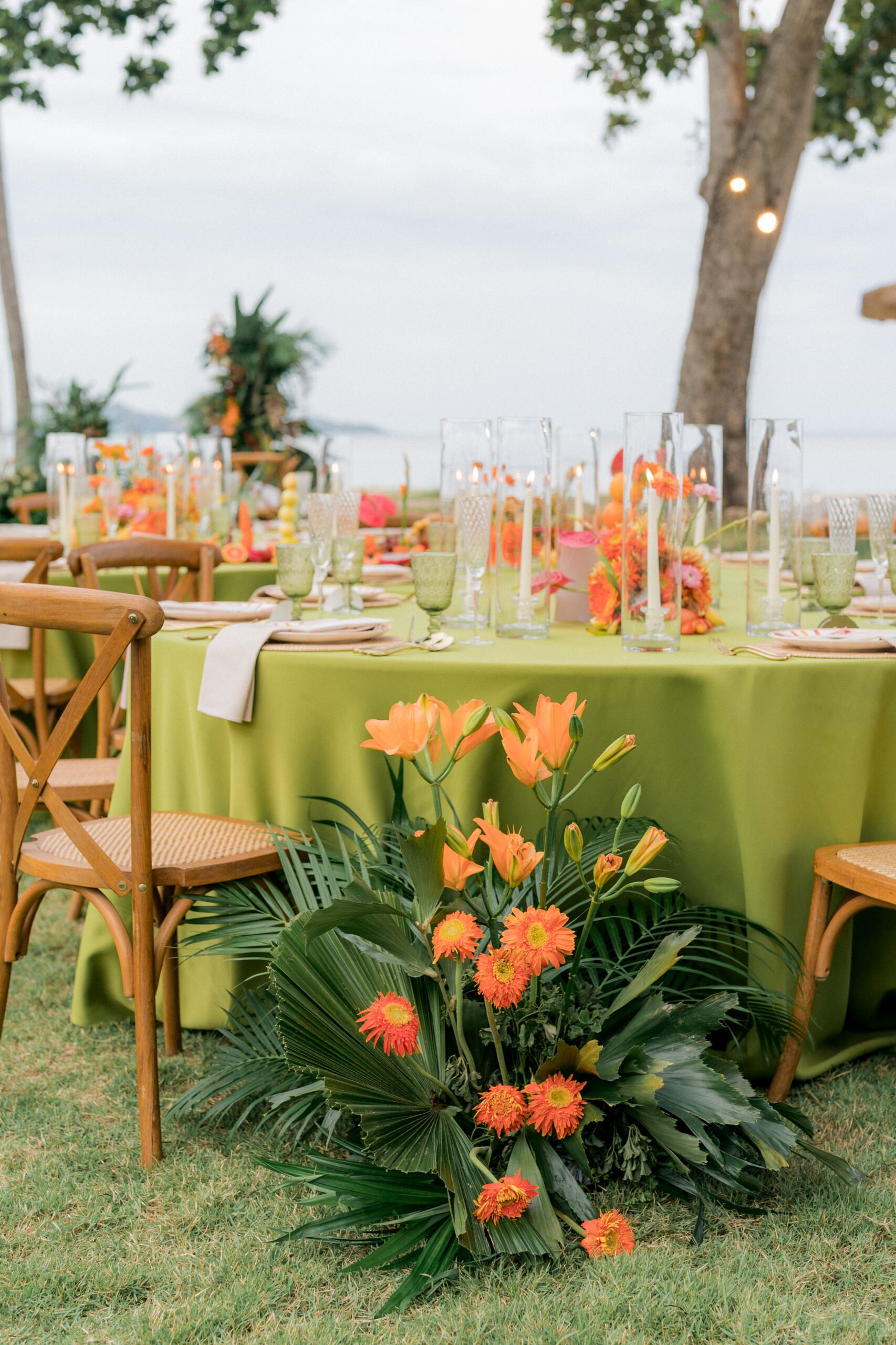 Outdoor table set for a celebration with a green tablecloth, surrounded by wooden chairs, decorated with orange and pink flowers, tall candles, and glassware, with trees and water visible in the background.