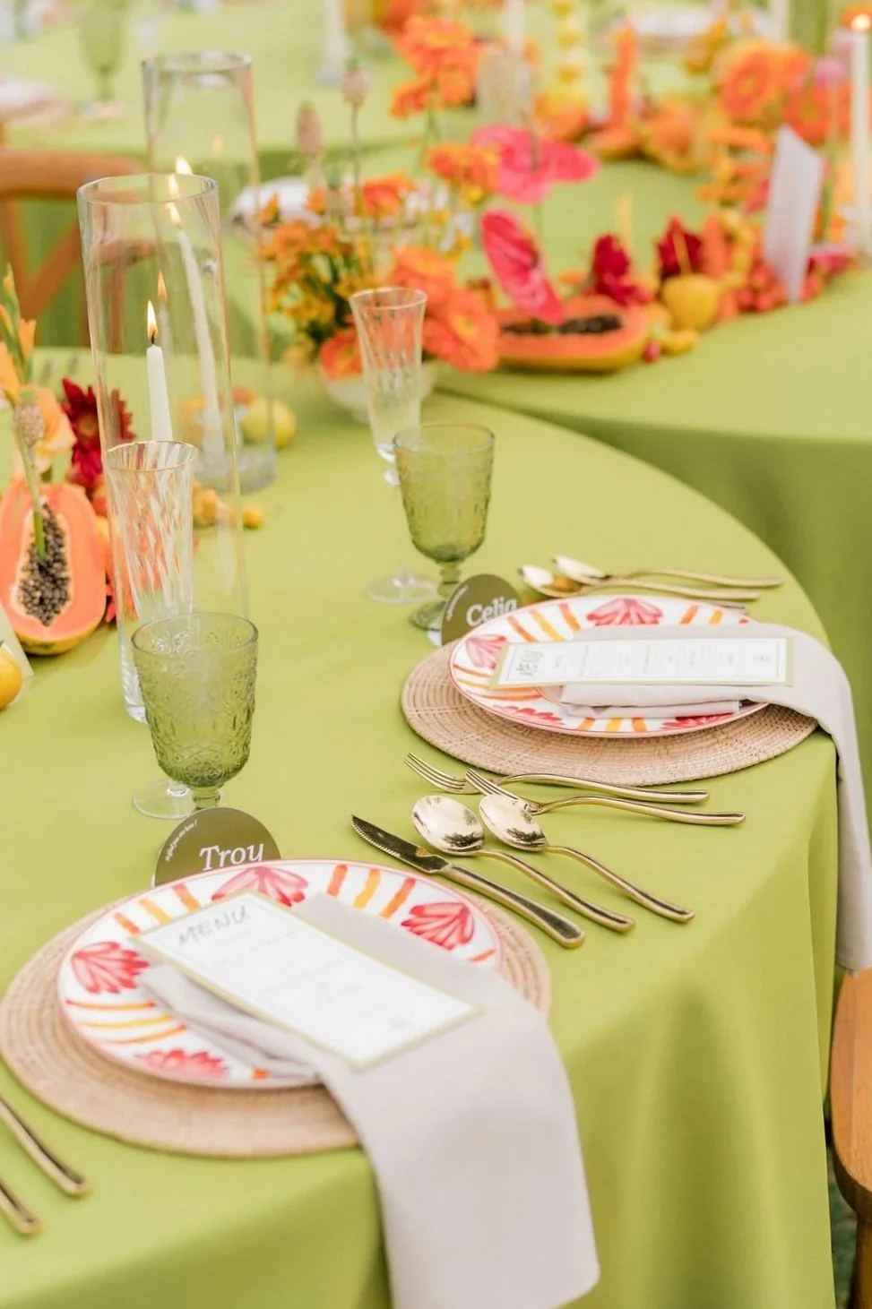 A brightly decorated table set for a tropical-themed event with green tablecloth, colorful flower arrangements, and candles. Each place setting includes a menu, patterned plate, pink and orange striped napkin, and gold cutlery.