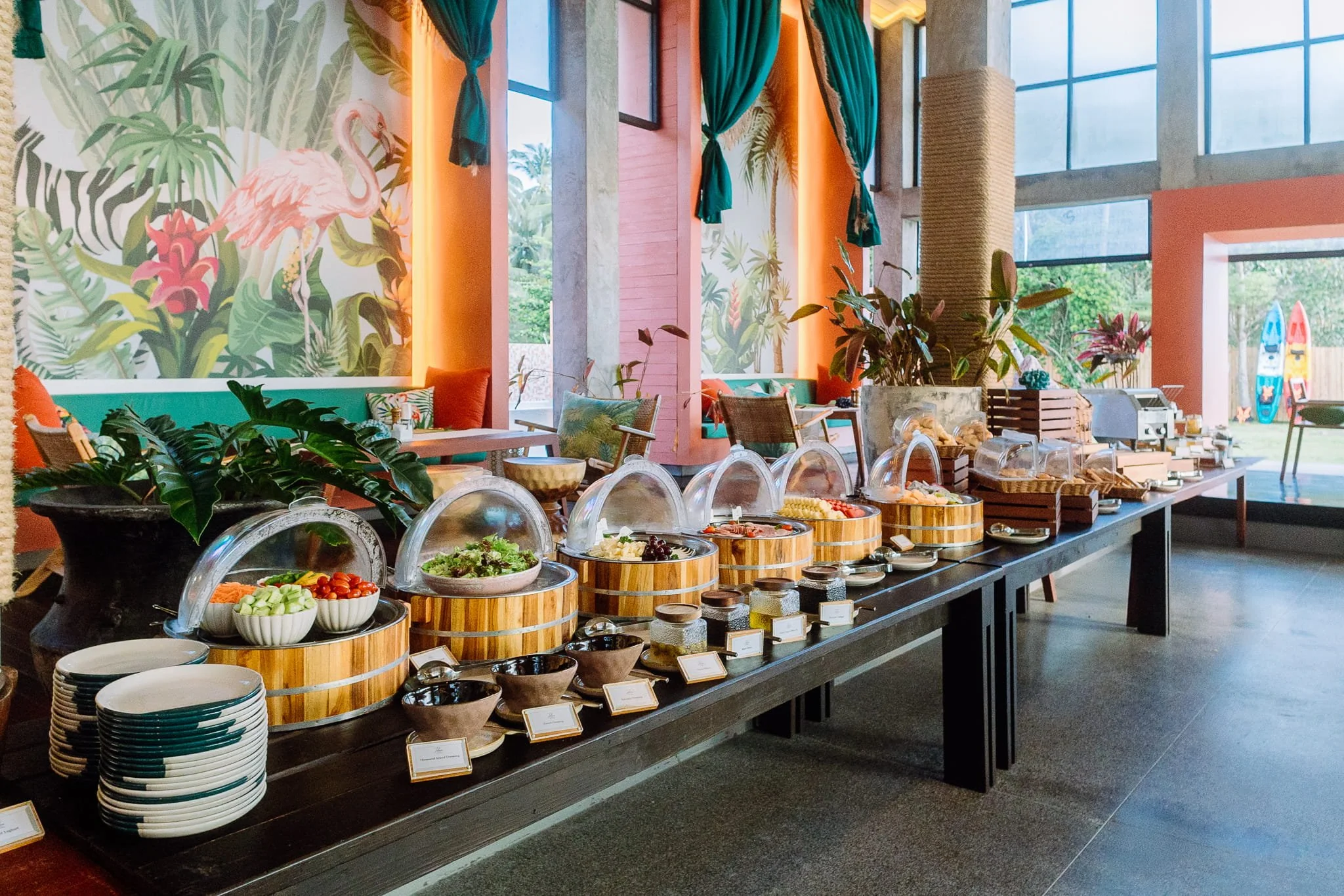 Buffet table with various salads and dishes in a bright, tropical-themed restaurant with large windows and colorful decor.