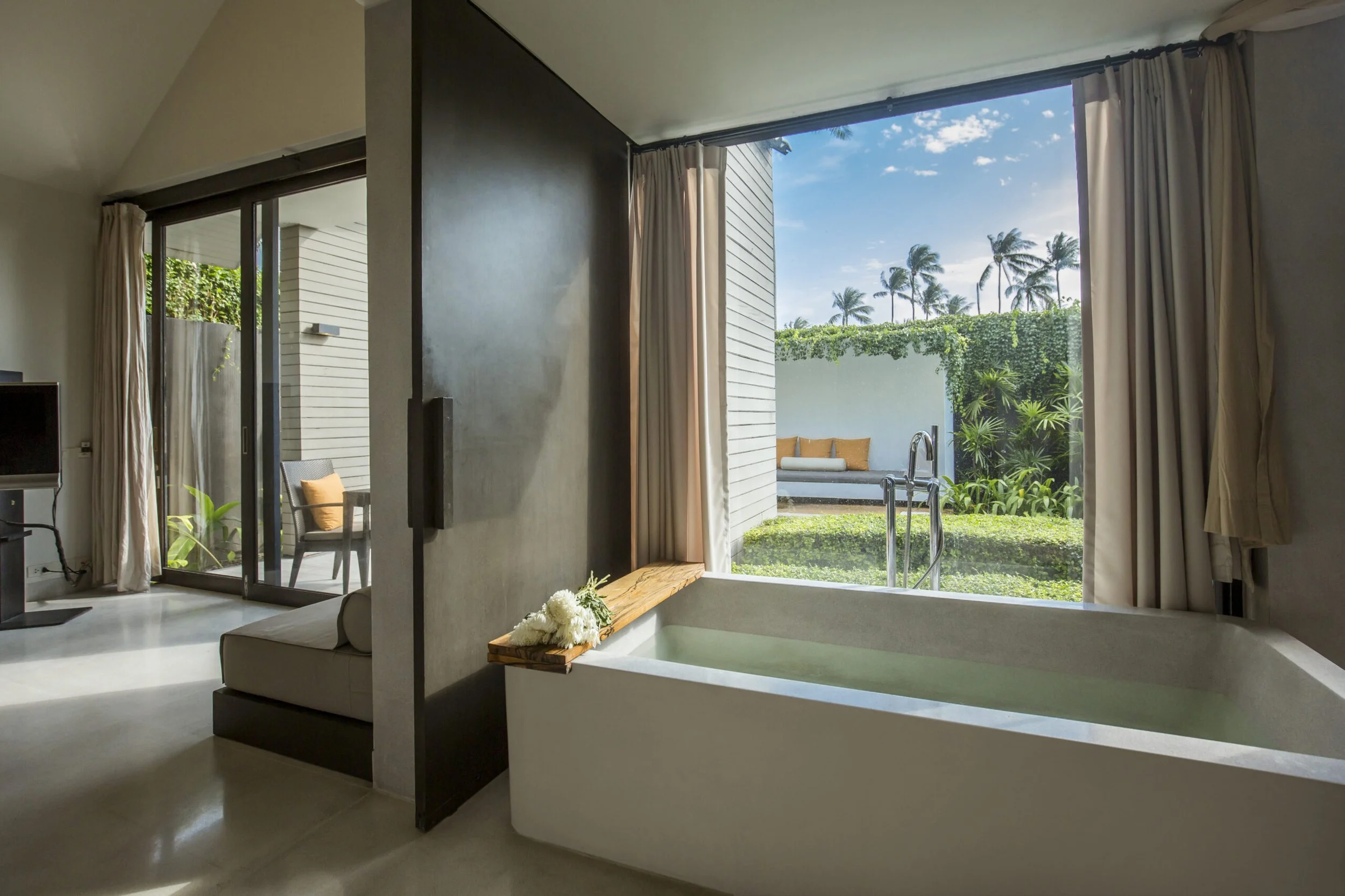 Hotel room with a bathtub next to a large open window overlooking a garden and patio with outdoor seating, tropical plants, and palm trees.
