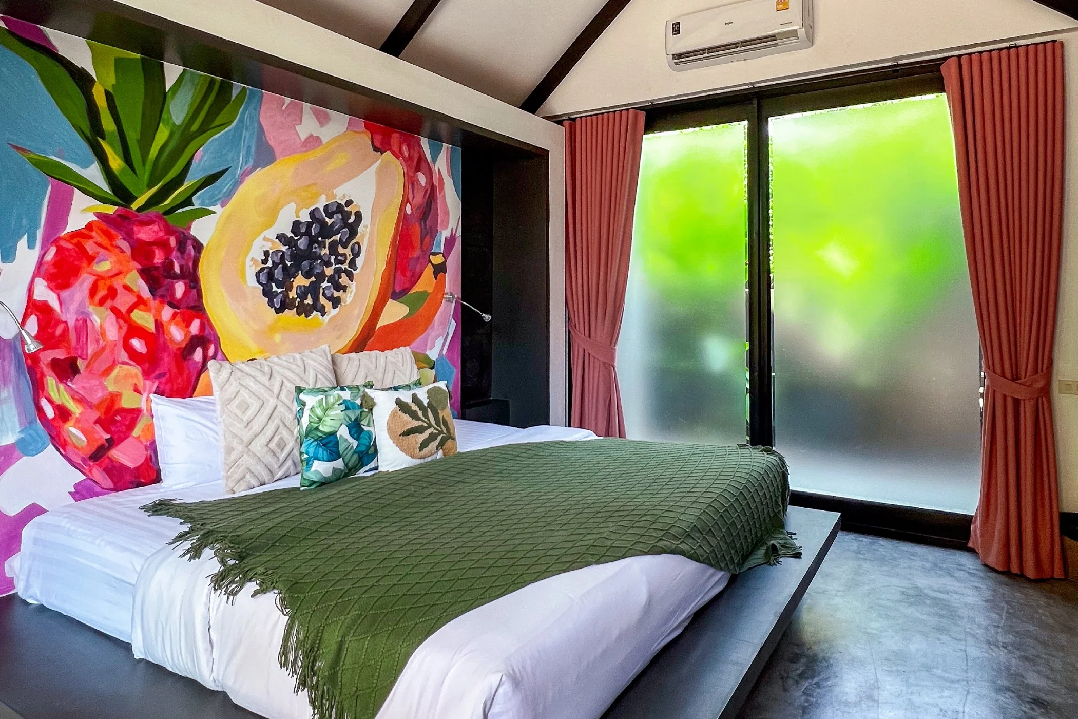 A bedroom with a vibrant fruit-themed mural on the wall behind the bed, featuring a pineapple, papaya, and strawberries. The bed is made with white linens, several decorative pillows, and a dark green quilted blanket. There are large frosted glass sliding doors with pink curtains, and an air conditioning unit mounted on the wall.