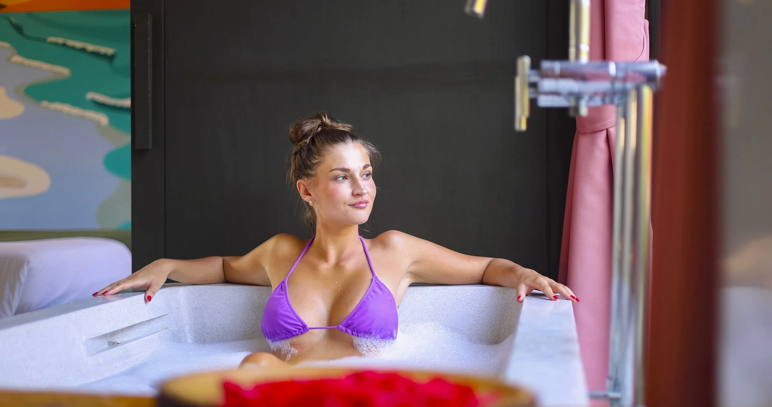 A young woman with a purple bikini top relaxing in a white bathtub filled with bubbles, looking out thoughtfully, with her arms resting on the sides of the tub.
