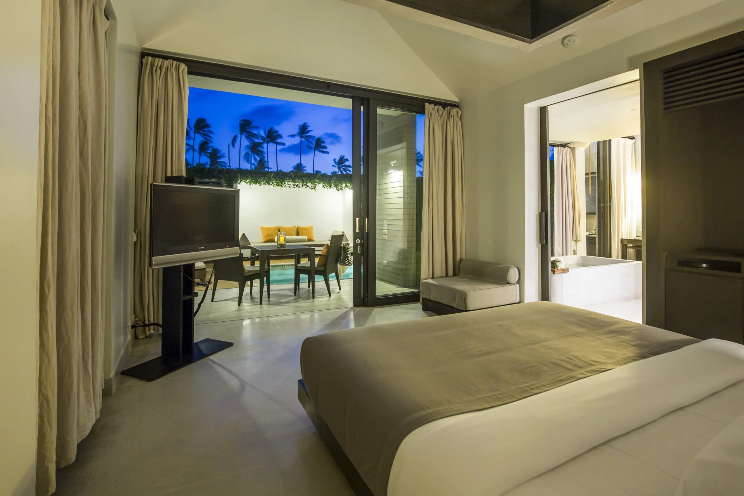 View of a hotel room with a bed, a television on a stand, and an open door leading to a balcony with outdoor dining furniture under palm trees at dusk.