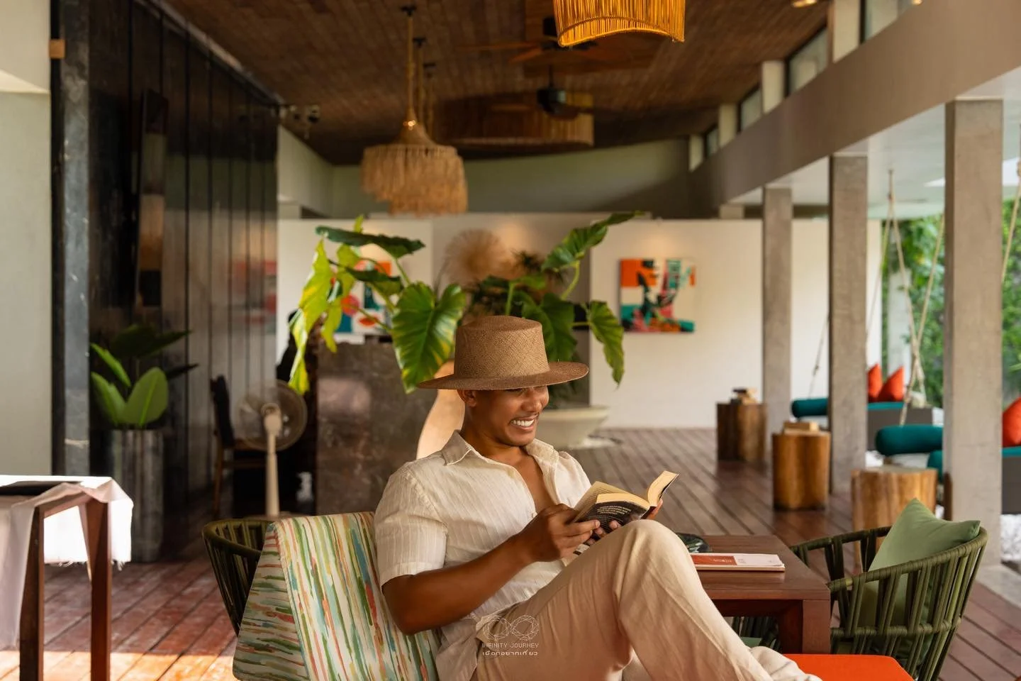 A woman sitting at a table indoors, smiling while reading a book. She is wearing a large straw hat and light-colored clothes, with a cozy and modern decor background.