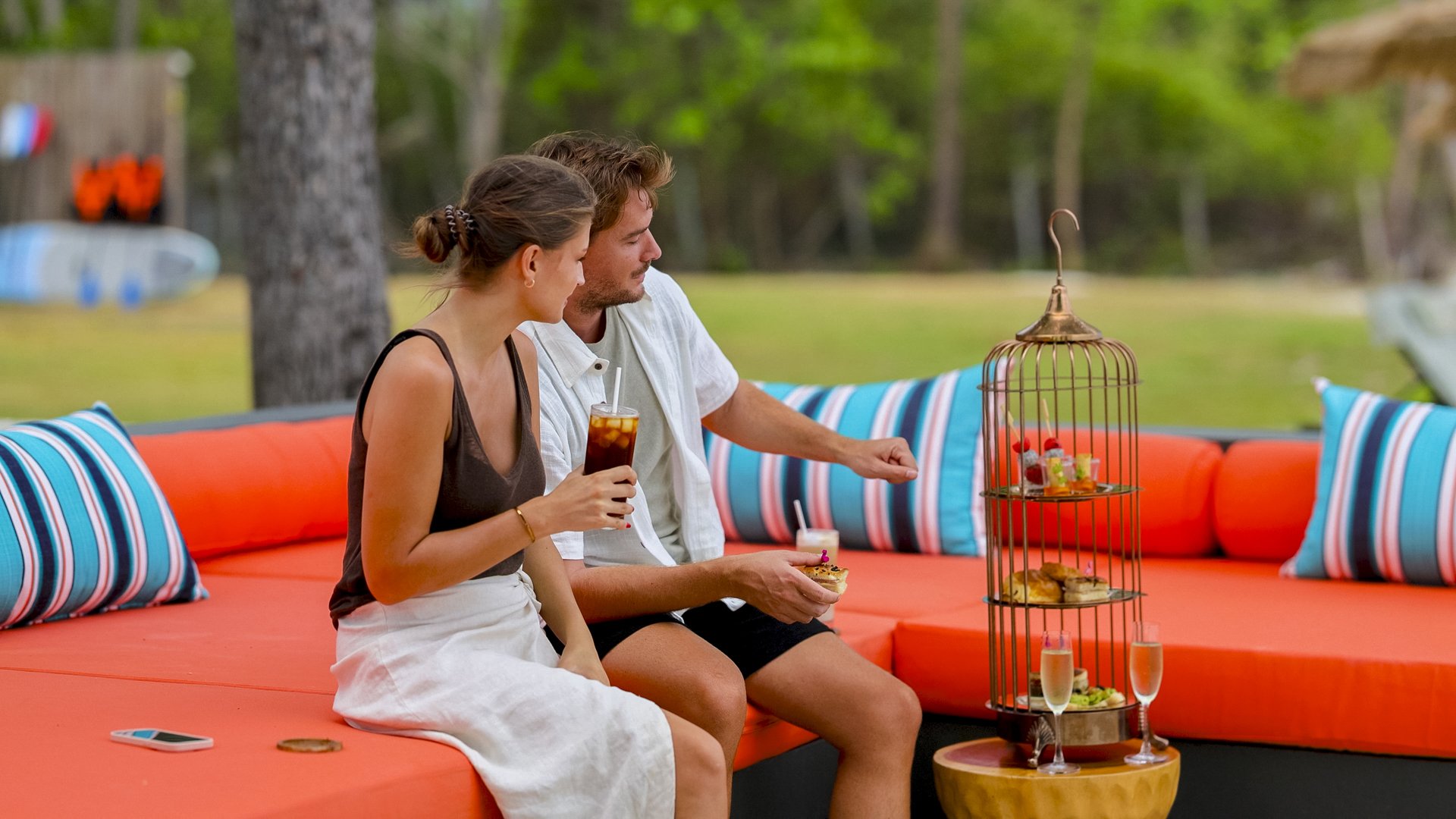 A young woman and man sitting on an outdoor orange sectional sofa with striped pillows, sharing a drink and snack, with a birdcage-style serving stand holding desserts and champagne glasses nearby, in a lush green backyard.