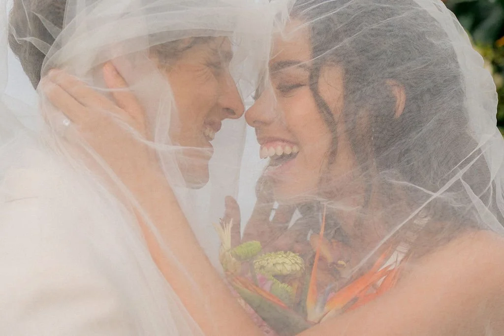 Two women embracing with their foreheads and noses touching, smiling through a sheer veil during a wedding or special celebration.