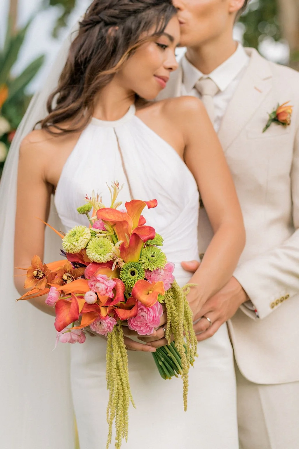 A woman in a white sleeveless wedding dress holding a colorful bouquet of orange, pink, and green flowers while standing close to a man in a cream suit. The man is kissing her forehead, and they appear to be at a wedding.