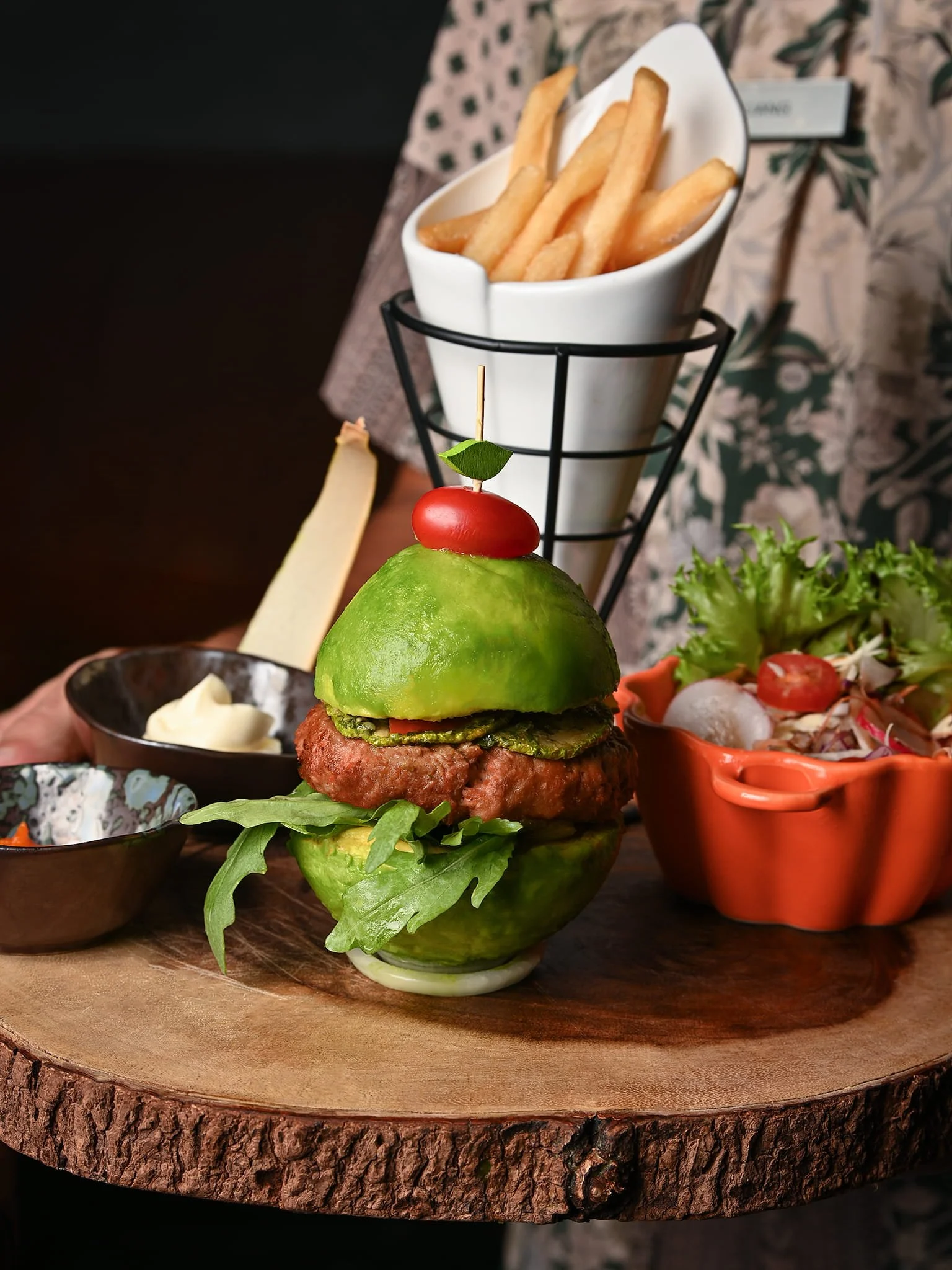 A burger decorated with a slice of avocado, cherry tomato, and a leafy herb on top, served with French fries in a white container, salad in an orange bowl, and small side dishes, all placed on a wooden serving board.