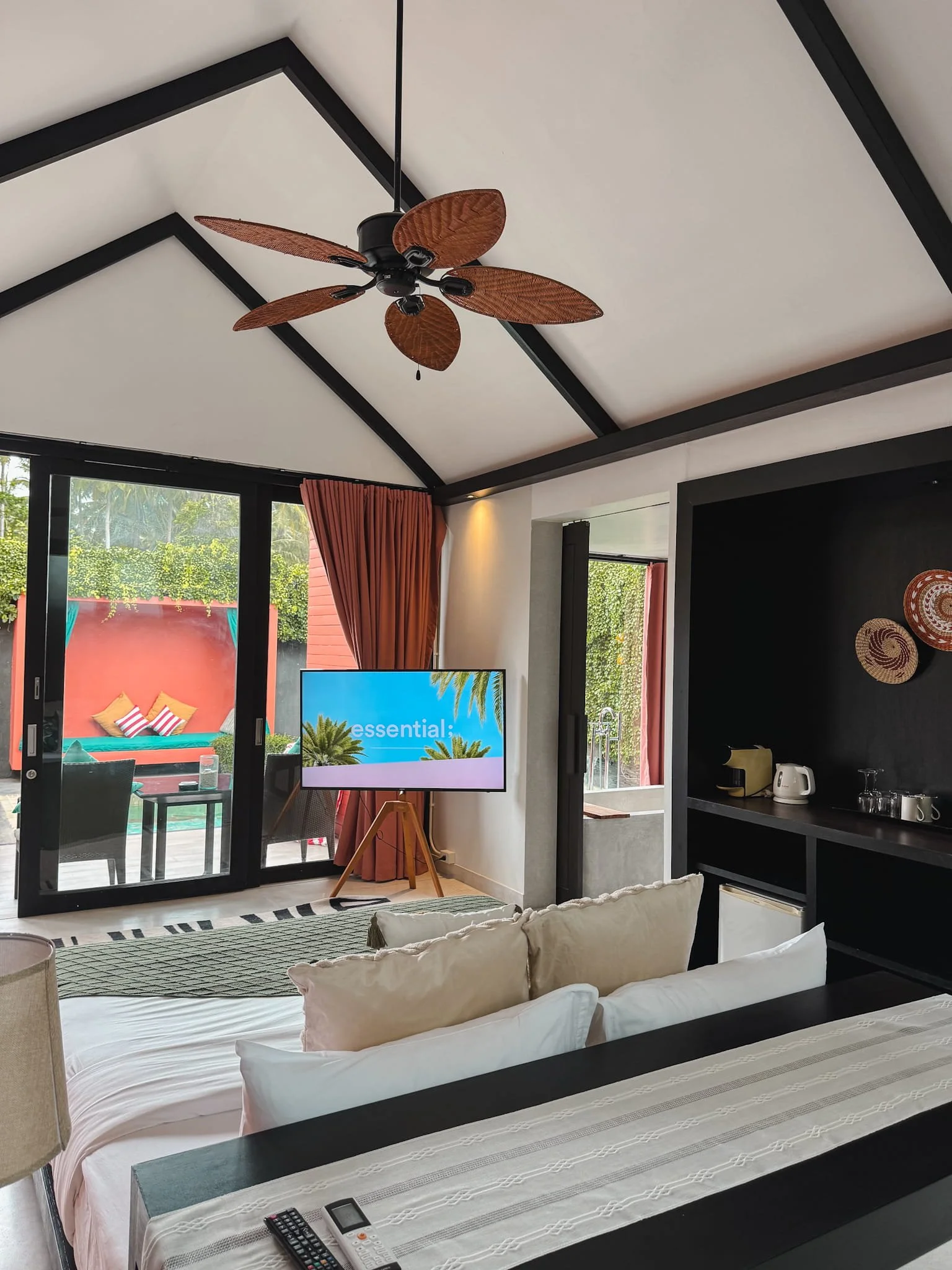 Interior of a modern hotel room with a ceiling fan, a flat-screen TV, a sliding glass door with curtains leading to an outdoor balcony, a bed with pillows, and a kitchenette area.