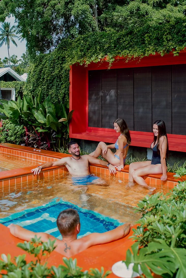 Group of young adults enjoying time in a backyard pool surrounded by lush greenery and tropical plants, with a modern red wall and black window in the background.